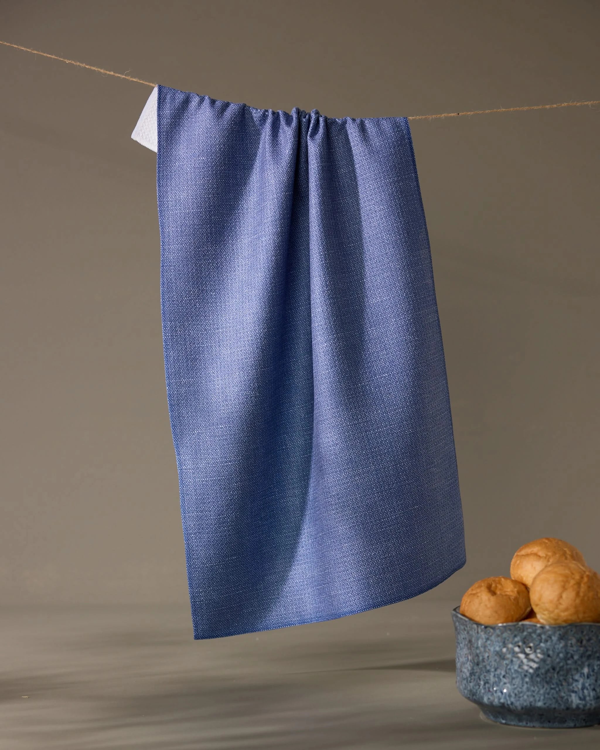 The Geometry Rainfield tea towel in textured blue hangs from a string against a plain background, with a ceramic bowl of three bread rolls placed below on the right.
