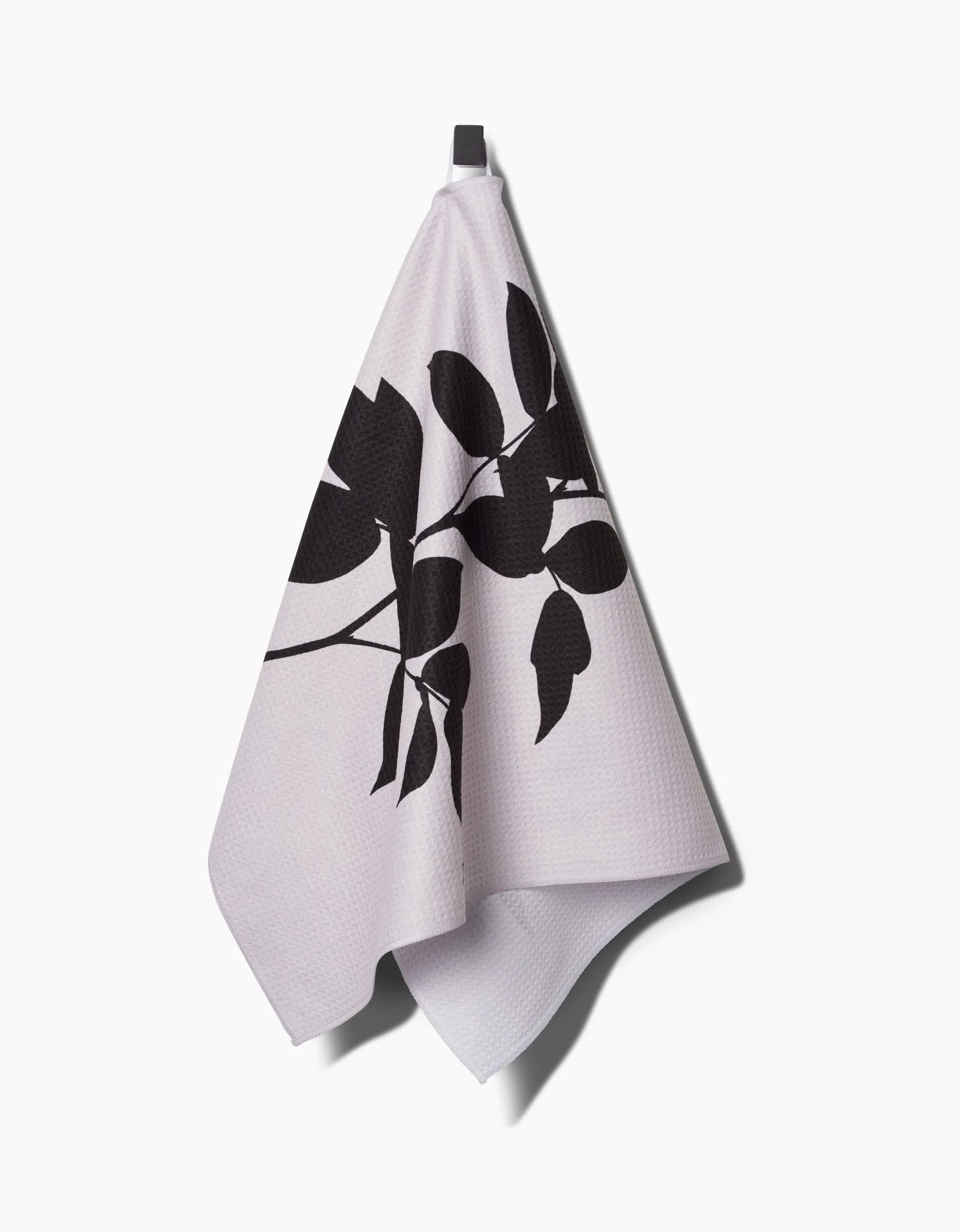A light-colored PrintedPlant Raspberry Tea Towel made from Geoweave™ fabric with a black botanical leaf pattern hangs from a hook against a white background.