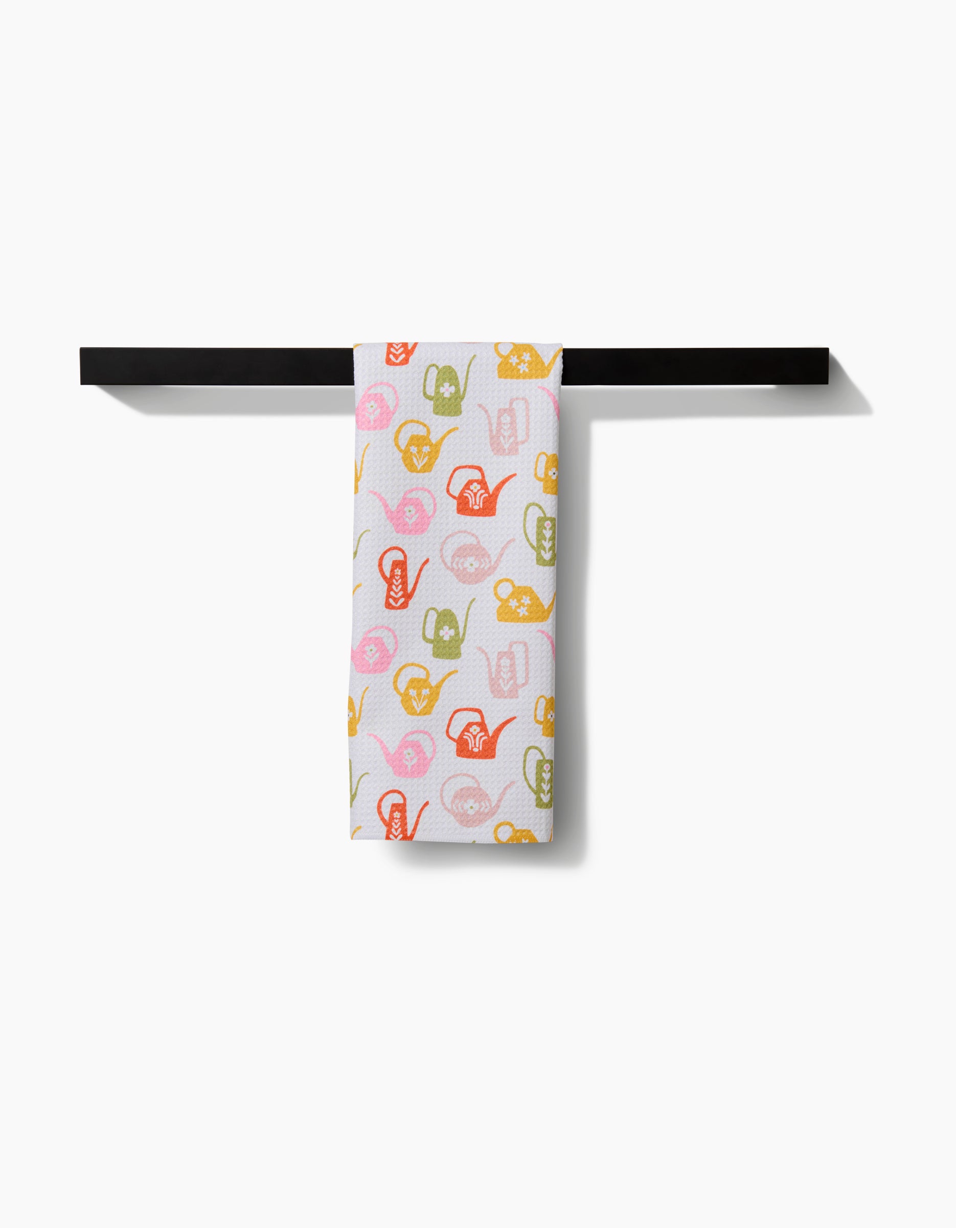 A Kayla Ann Retro Watering Cans tea towel featuring vibrant watering can designs hangs neatly on a simple black horizontal bar against a plain white background.
