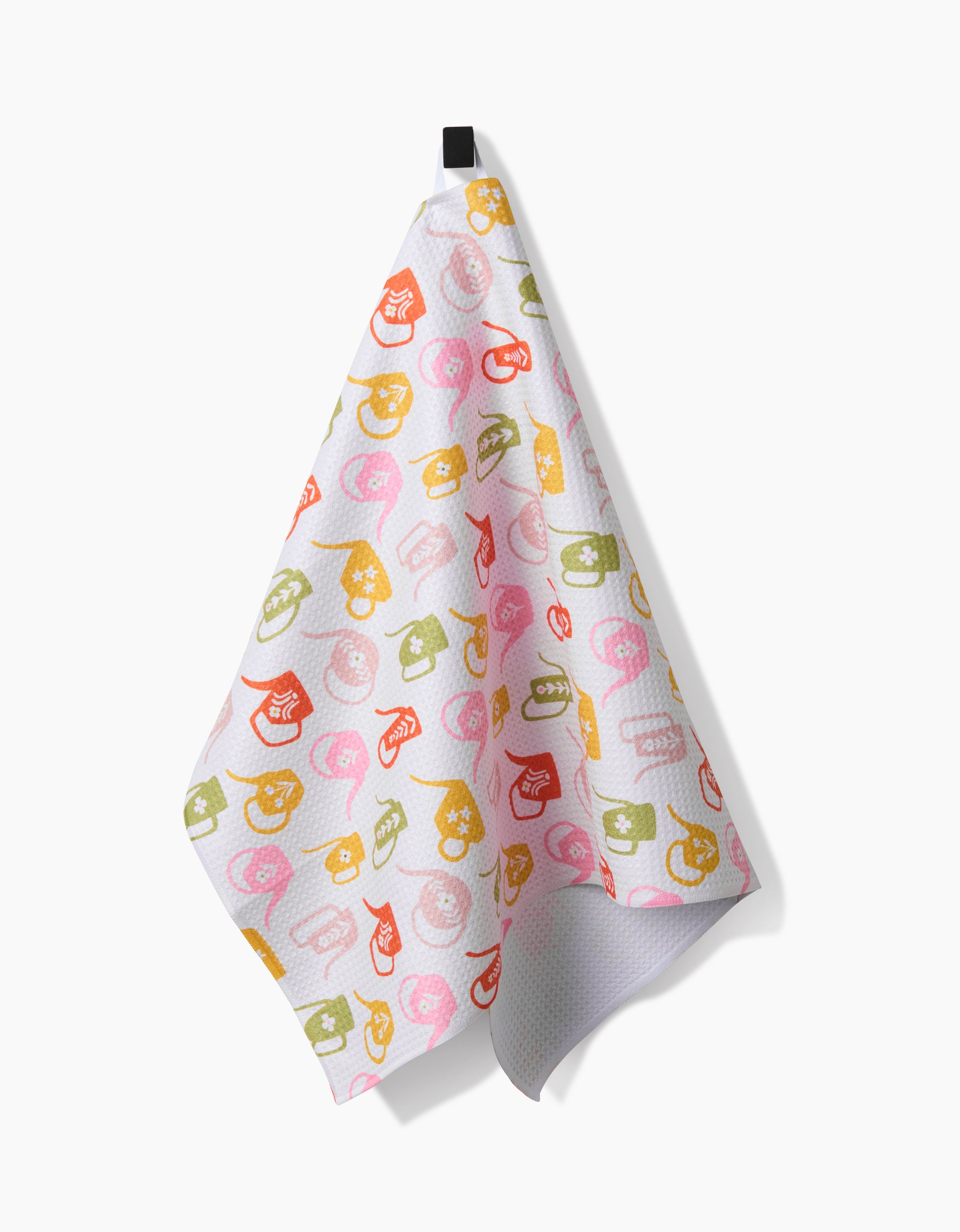 A white Kayla Ann Retro Watering Cans tea towel, featuring colorful watering cans on Geoweave fabric, hangs on a black hook against a plain white background.