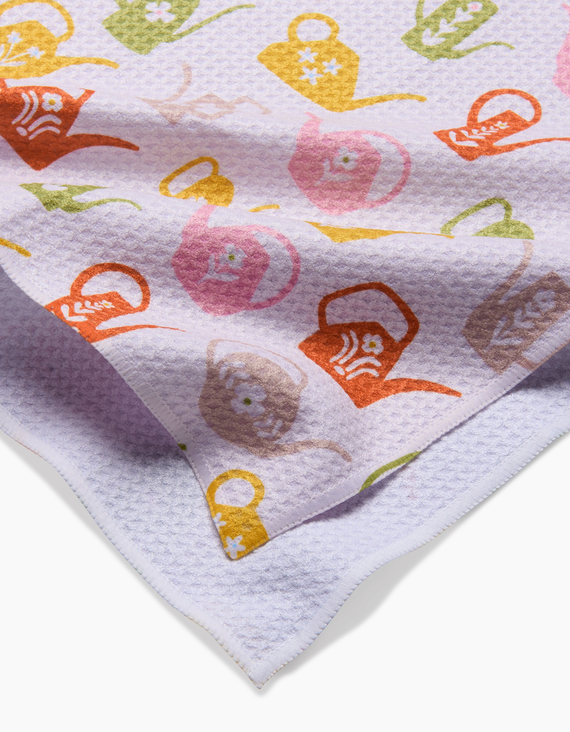 A close-up of a Kayla Ann Retro Watering Cans tea towel made from Geoweave fabric shows its textured white surface and colorful orange, yellow, pink, and green pattern. The folded towel highlights the vibrant design on both layers.