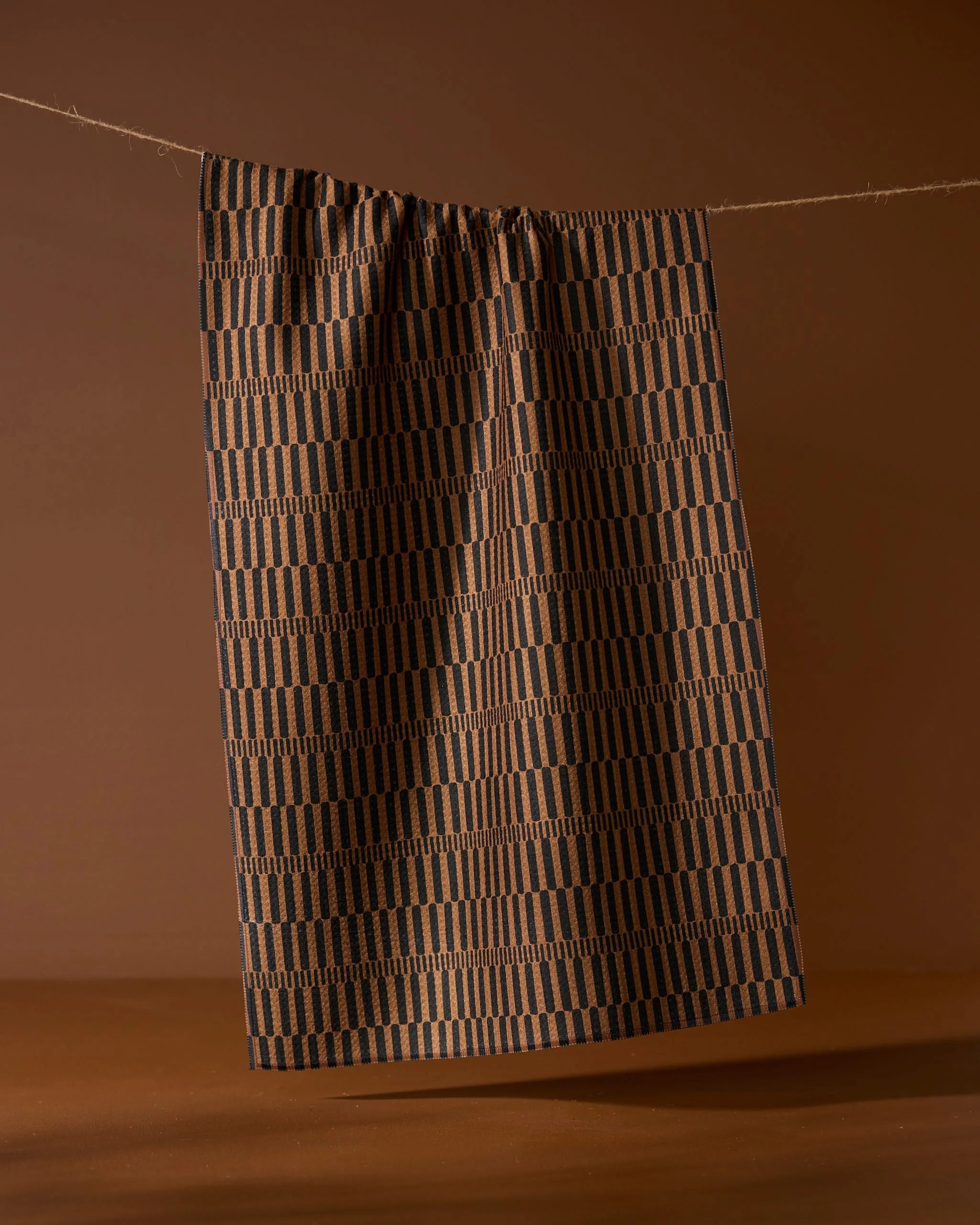 The Geometry Ridge tea towel, featuring a brown and black geometric pattern, hangs from a string against a brown background.