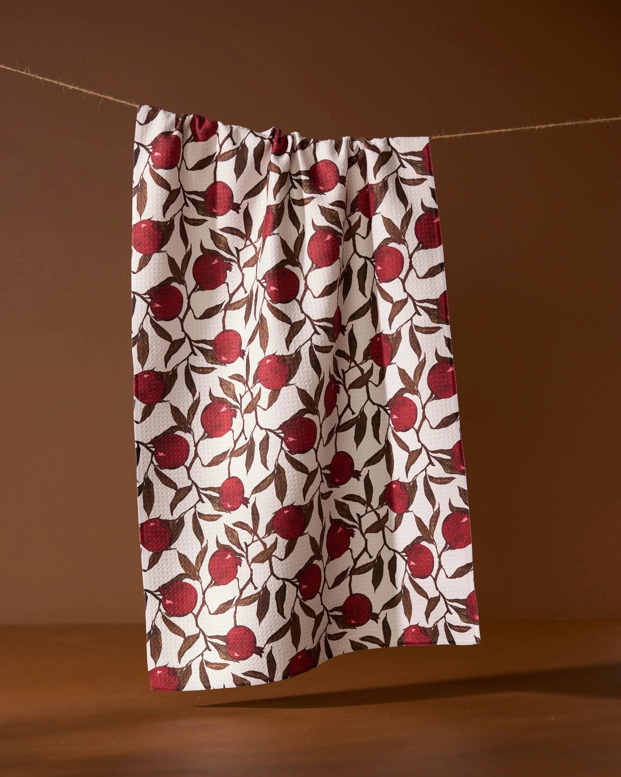The Geometry Rind tea towel, featuring a red apple and green leaf pattern, hangs on a string against a brown background.
