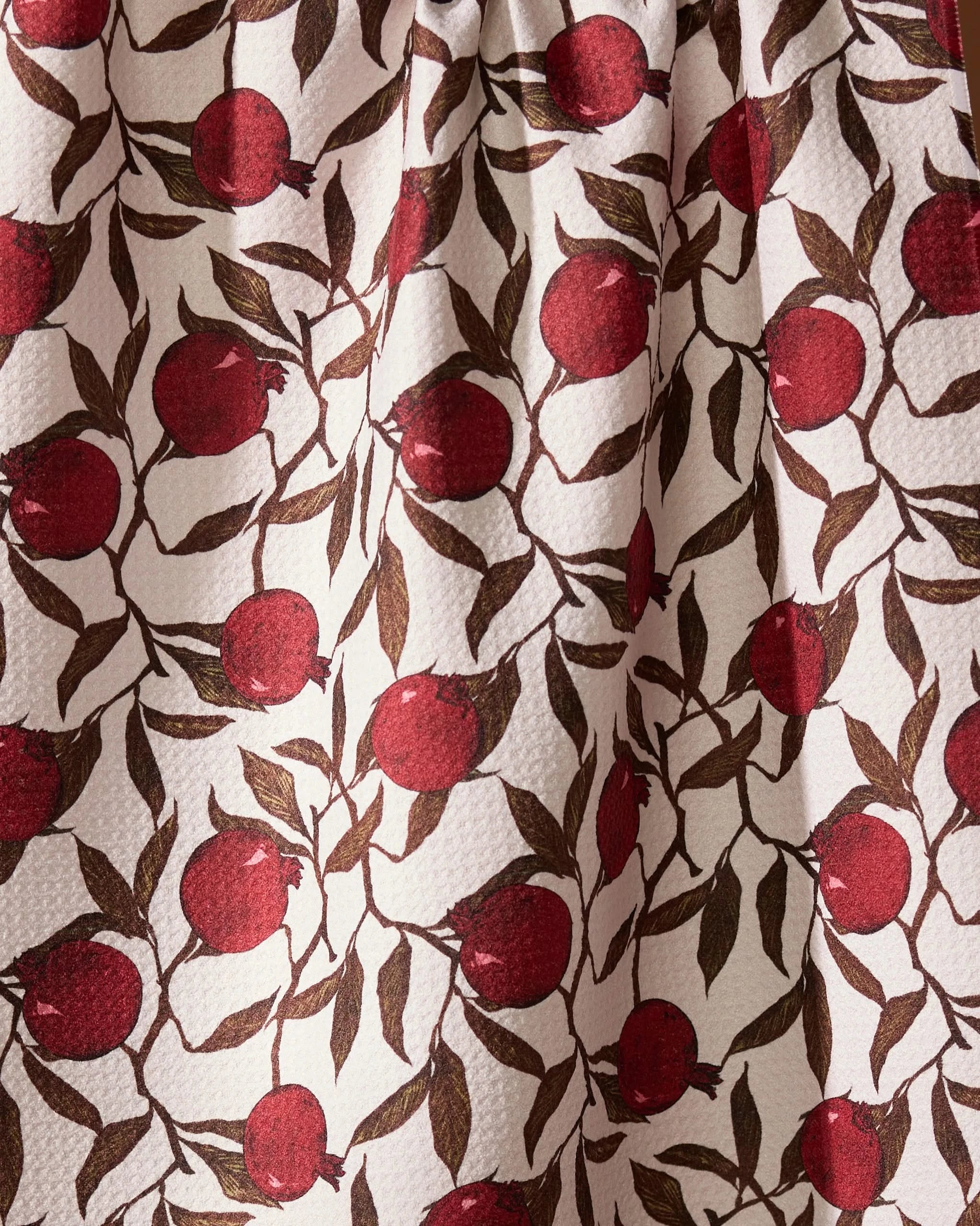 A close-up of the Geometry Rind tea towel, ultra-absorbent with a textured cream background, featuring a repeating pattern of red pomegranates and brown leafy branches.