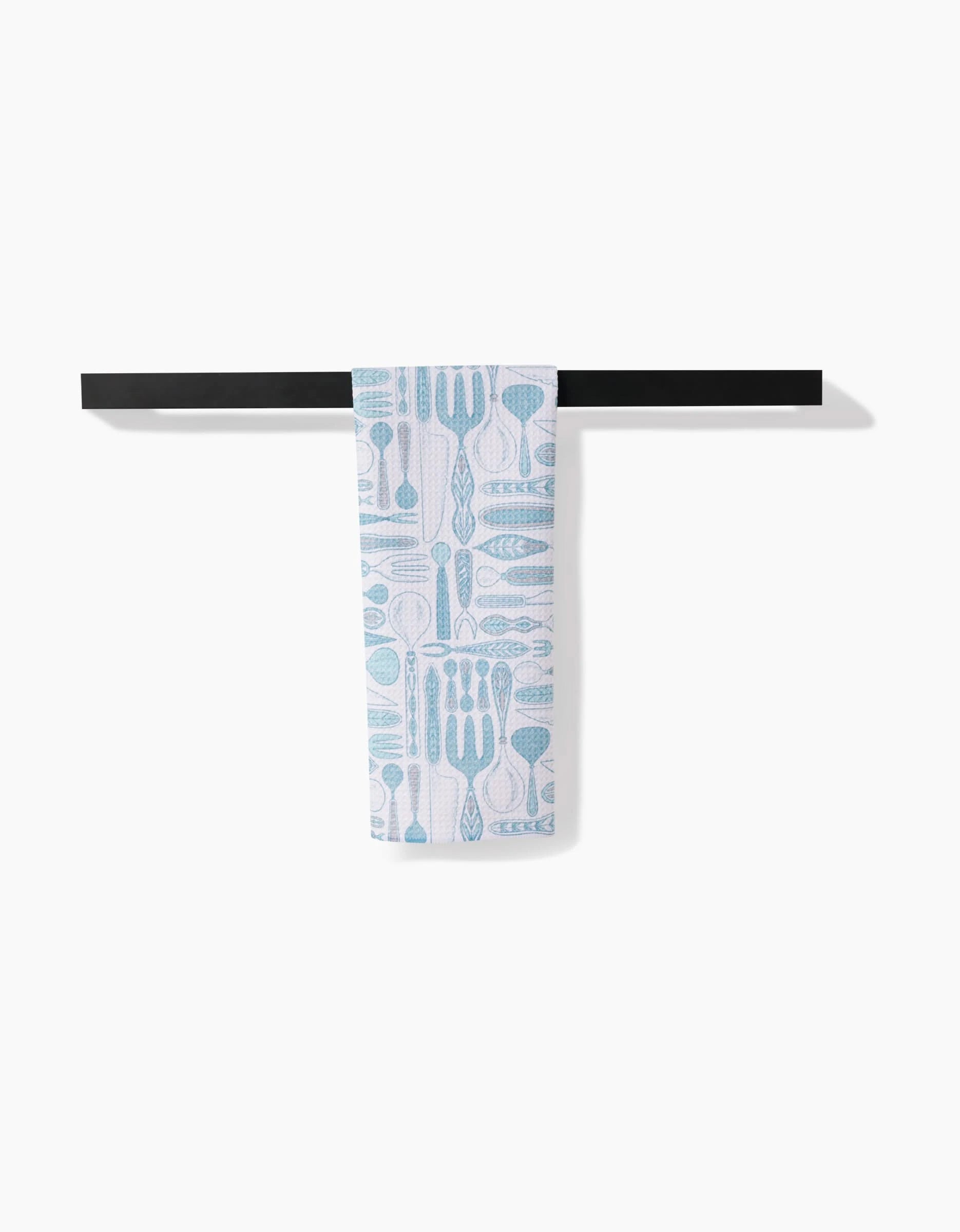 The Romy tea towel by Amy MacCready, featuring blue utensil patterns on ultra-absorbent white fabric, hangs neatly over a black horizontal towel bar against a plain white background.