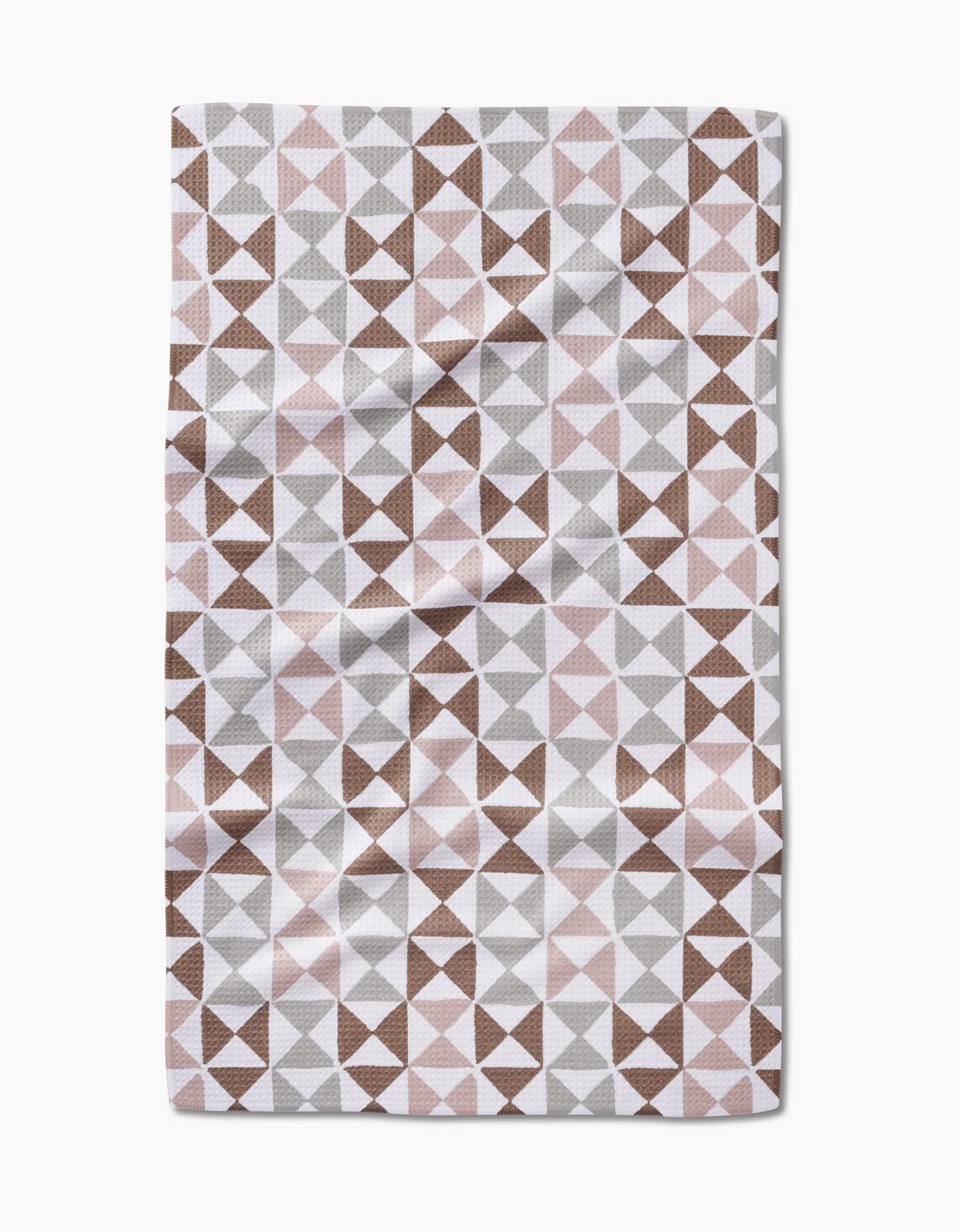 The Rubix tea towel by Studio Jeannot features a geometric triangle pattern in brown, gray, and light pink on Geoweave fabric made from recycled materials, shown with subtle creases against a white background.