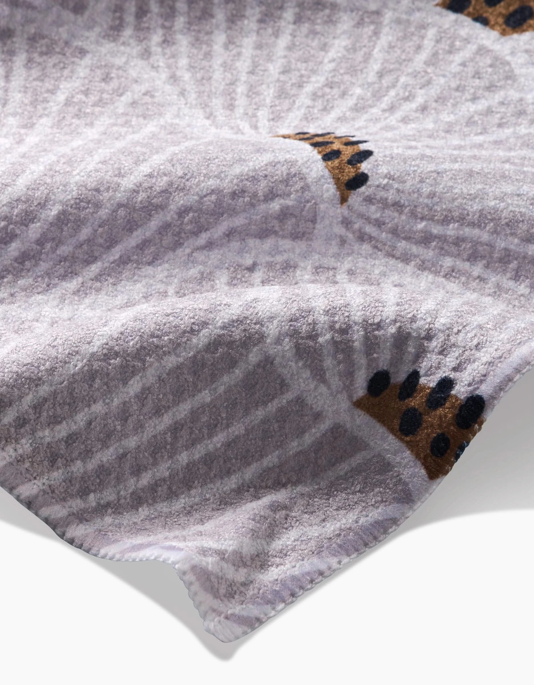 A close-up of the San Diego Tea Towel by Amy MacCready shows its soft, light purple Geoweave™ fabric with a subtle spiral pattern, brown spots with black dots like an animal print, and neatly stitched edges.