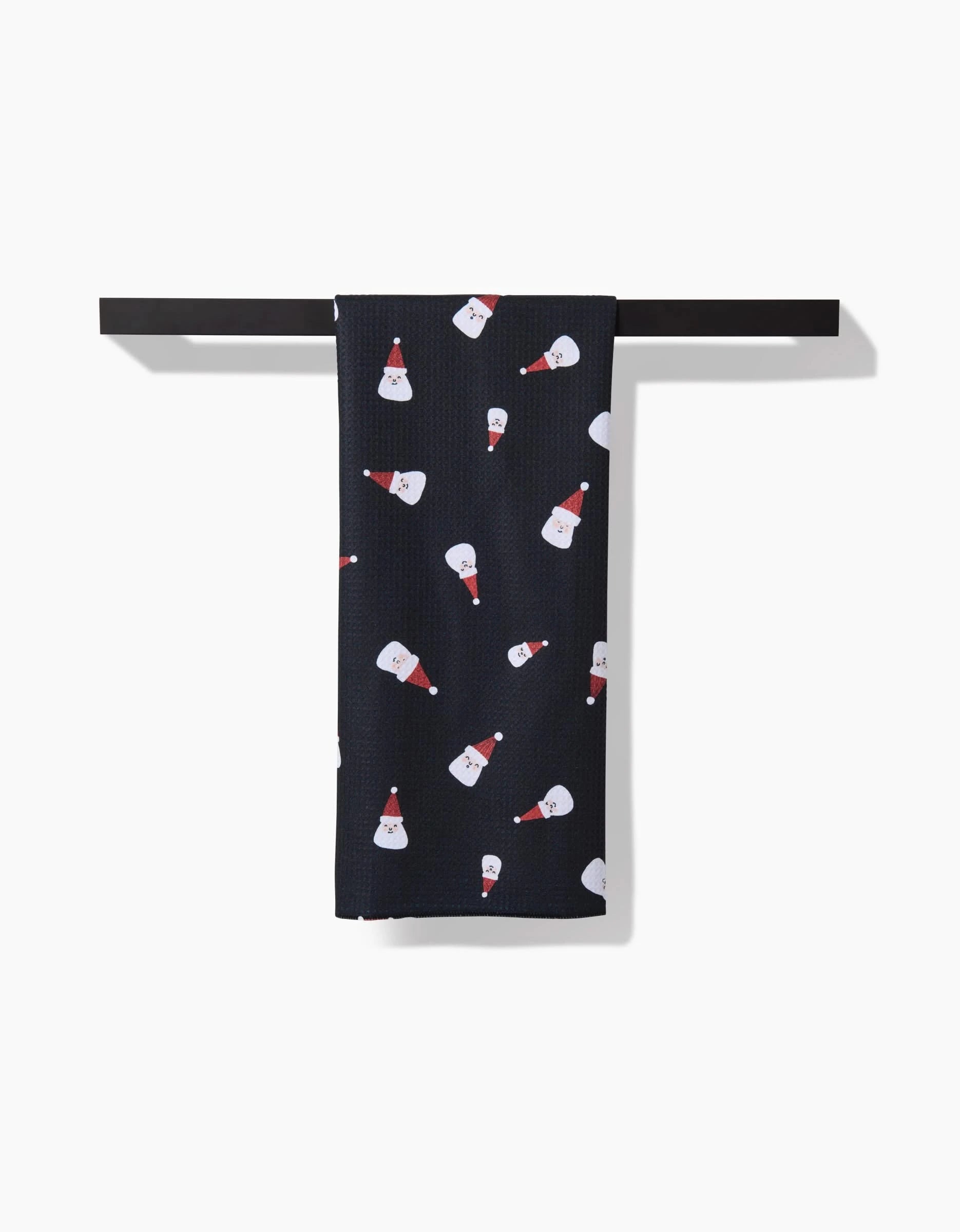 A black recycled Santa Faces kitchen towel by Coit Creative, featuring a pattern of smiling Santas in red hats, hangs neatly on a horizontal bar against a white background.