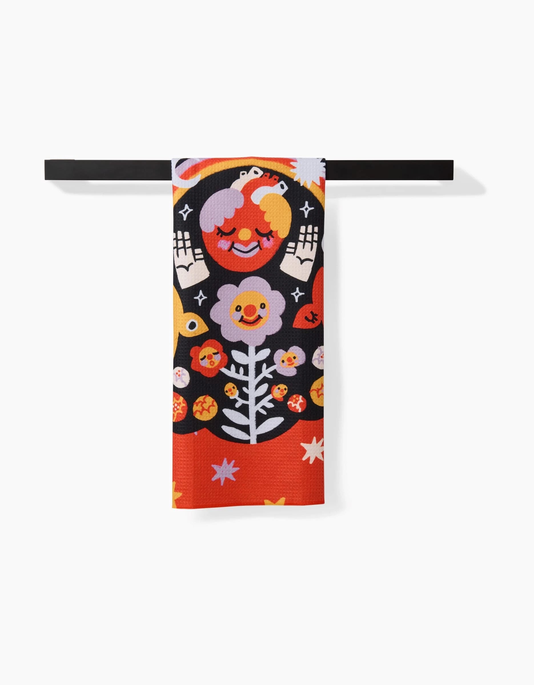 The Santa Muerte tea towel by So Lazo hangs on a black bar, displaying whimsical smiling faces, flowers, stars, and hands in bold orange, red, yellow, black, and white tones.