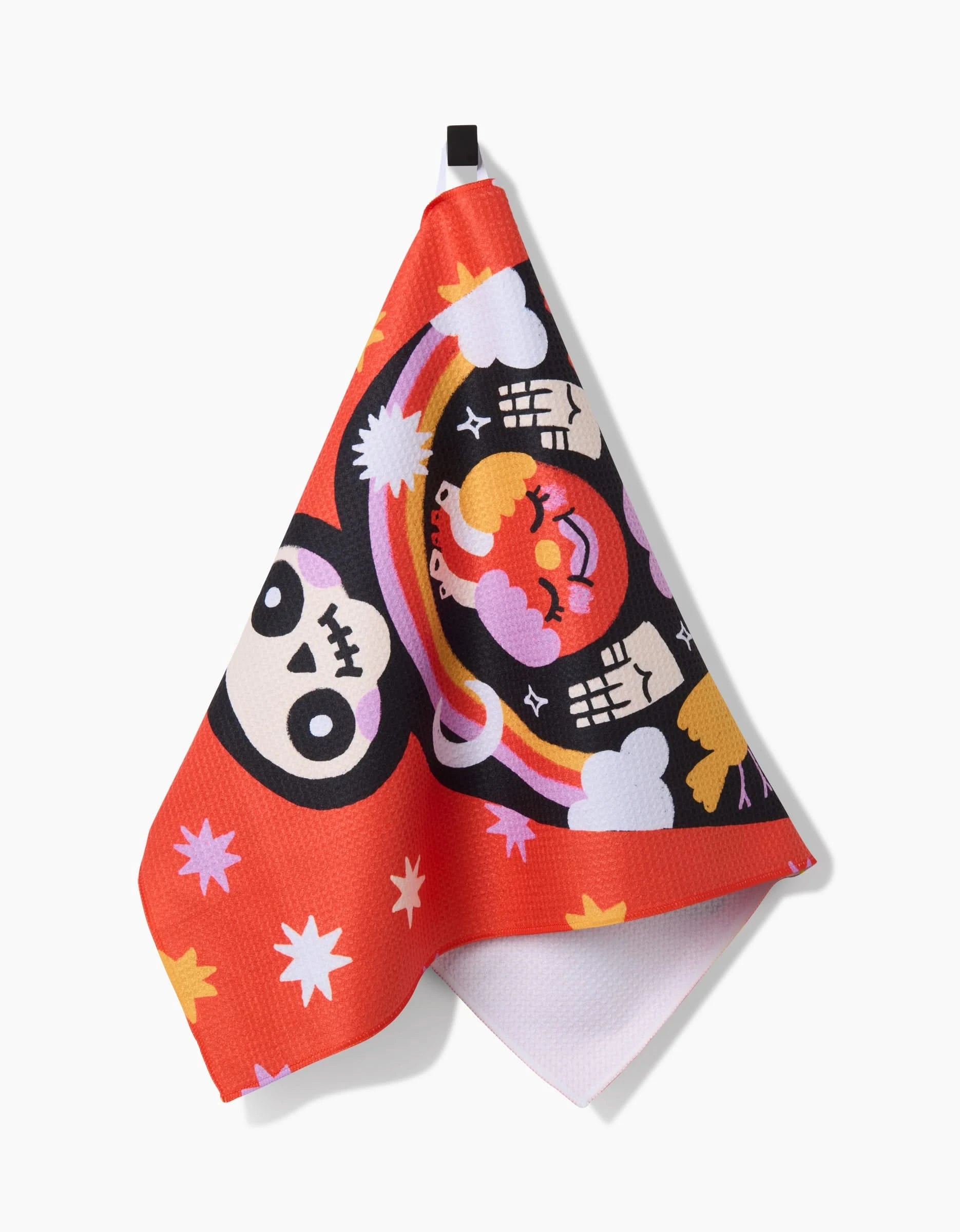 The Santa Muerte towel by So Lazo, featuring quick-dry fabric and vibrant cartoon skull faces, hands, stars, clouds, and a rainbow on red fabric, hangs from a black loop against a white background.