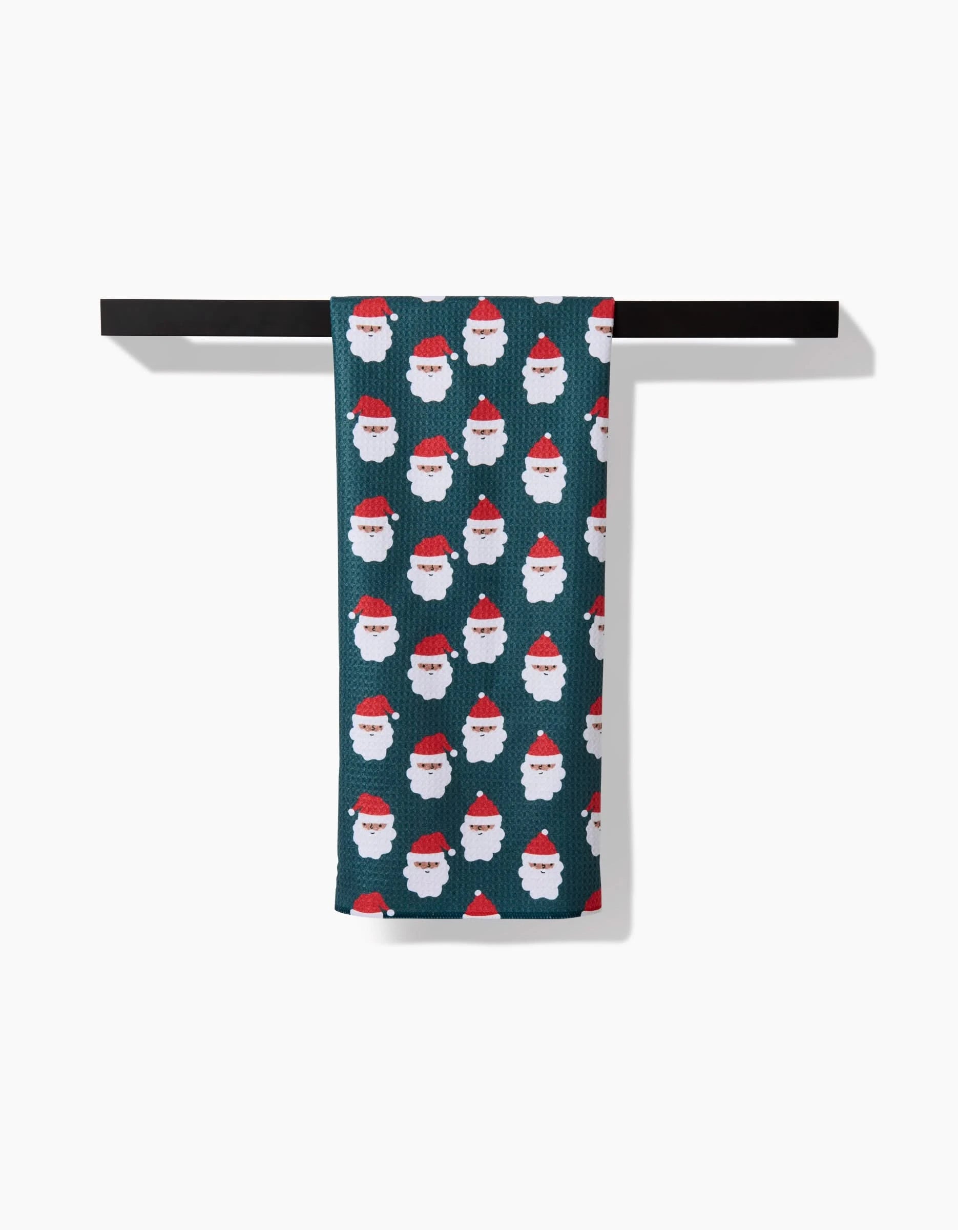 The Santa Santa kitchen towel by Charlie Rowan Designs, made from recycled materials, displays white Santas with red hats on dark green and hangs neatly on a black towel bar against a white background.