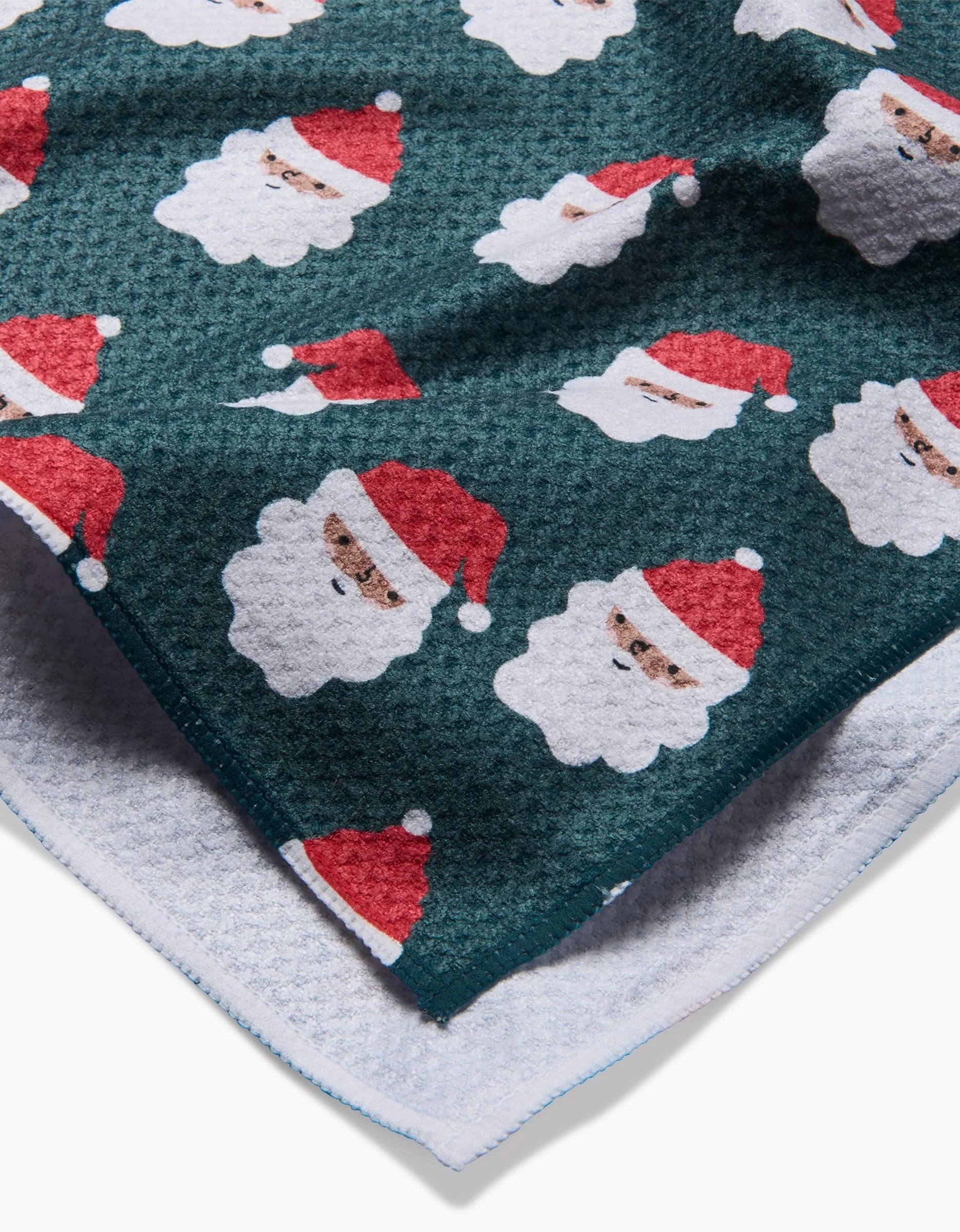 A close-up of the Charlie Rowan Designs Santa Santa tea towel shows its textured dark green fabric with repeating Santa faces in red hats and a soft white underside—great for festive cheer in your kitchen.