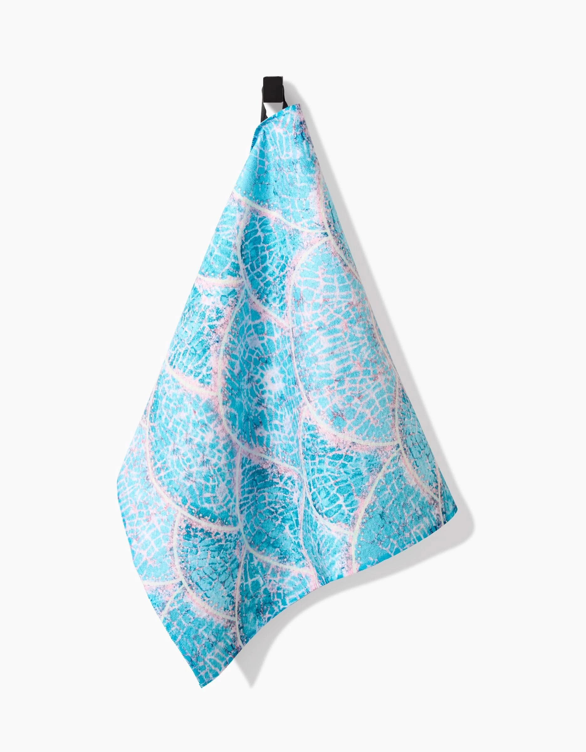 The Scallop Hills tea towel by Taryn Brennan, made from recycled materials and featuring a pool tile-inspired pattern in blue, hangs from a black loop against a plain white background.