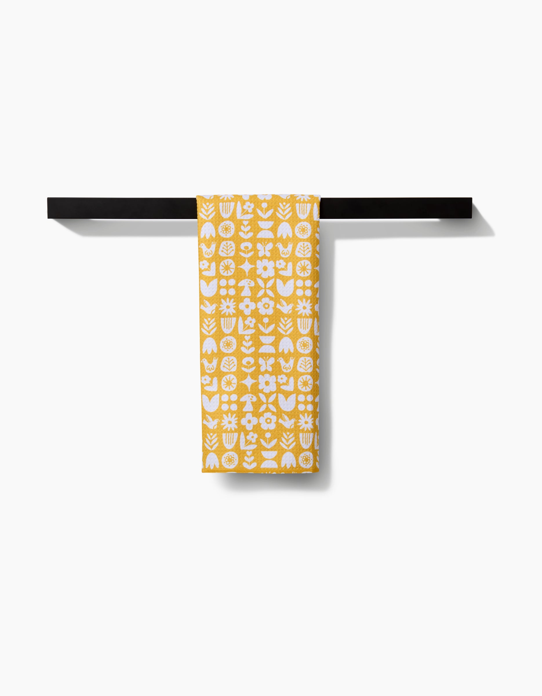 The Kayla Ann Scandi Spring Tea Towel, featuring yellow and white floral and geometric patterns on Geoweave fabric, hangs neatly on a black towel bar against a white background.