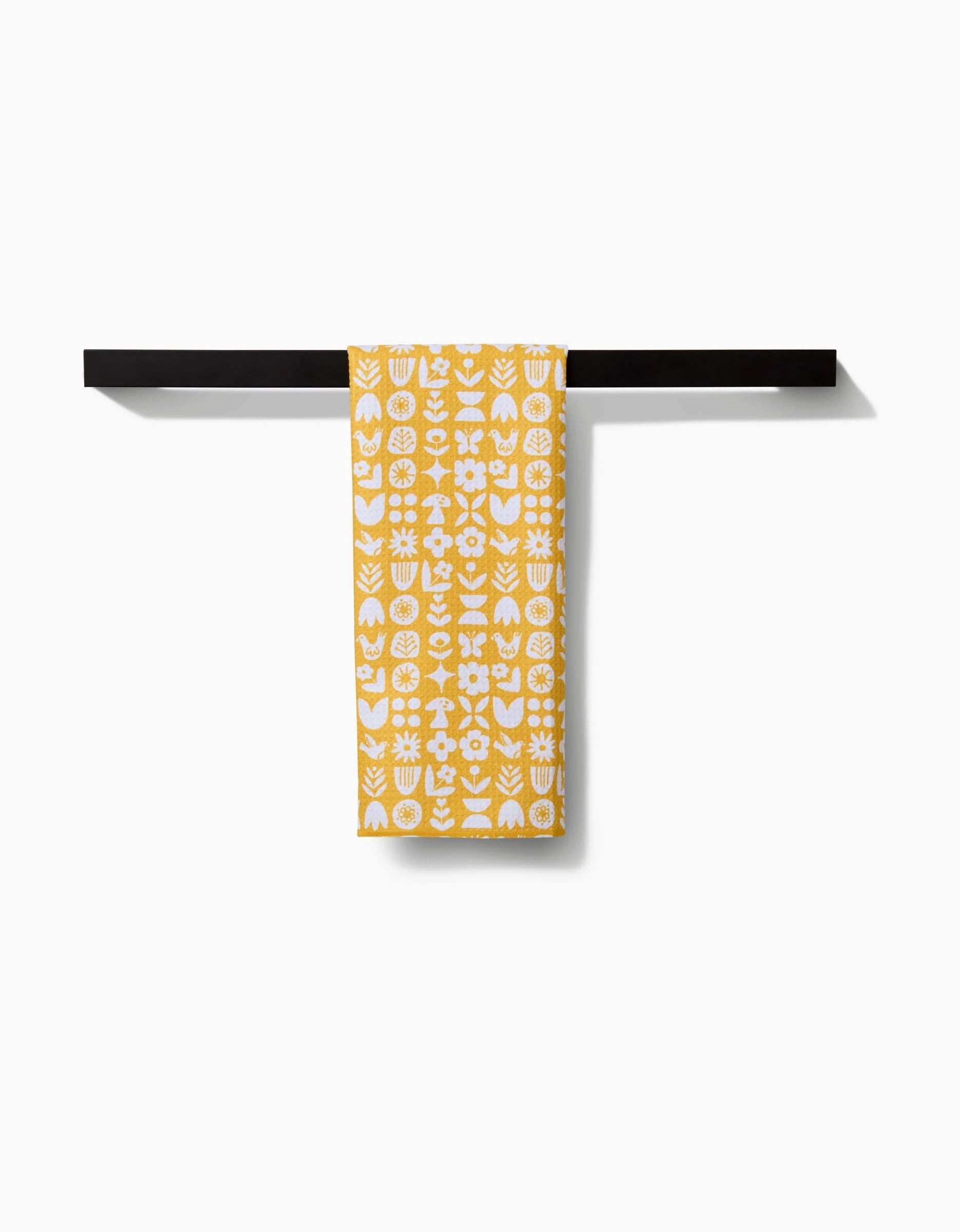 The Kayla Ann Scandi Spring Tea Towel, featuring yellow and white floral and geometric patterns on Geoweave fabric, hangs neatly on a black towel bar against a white background.