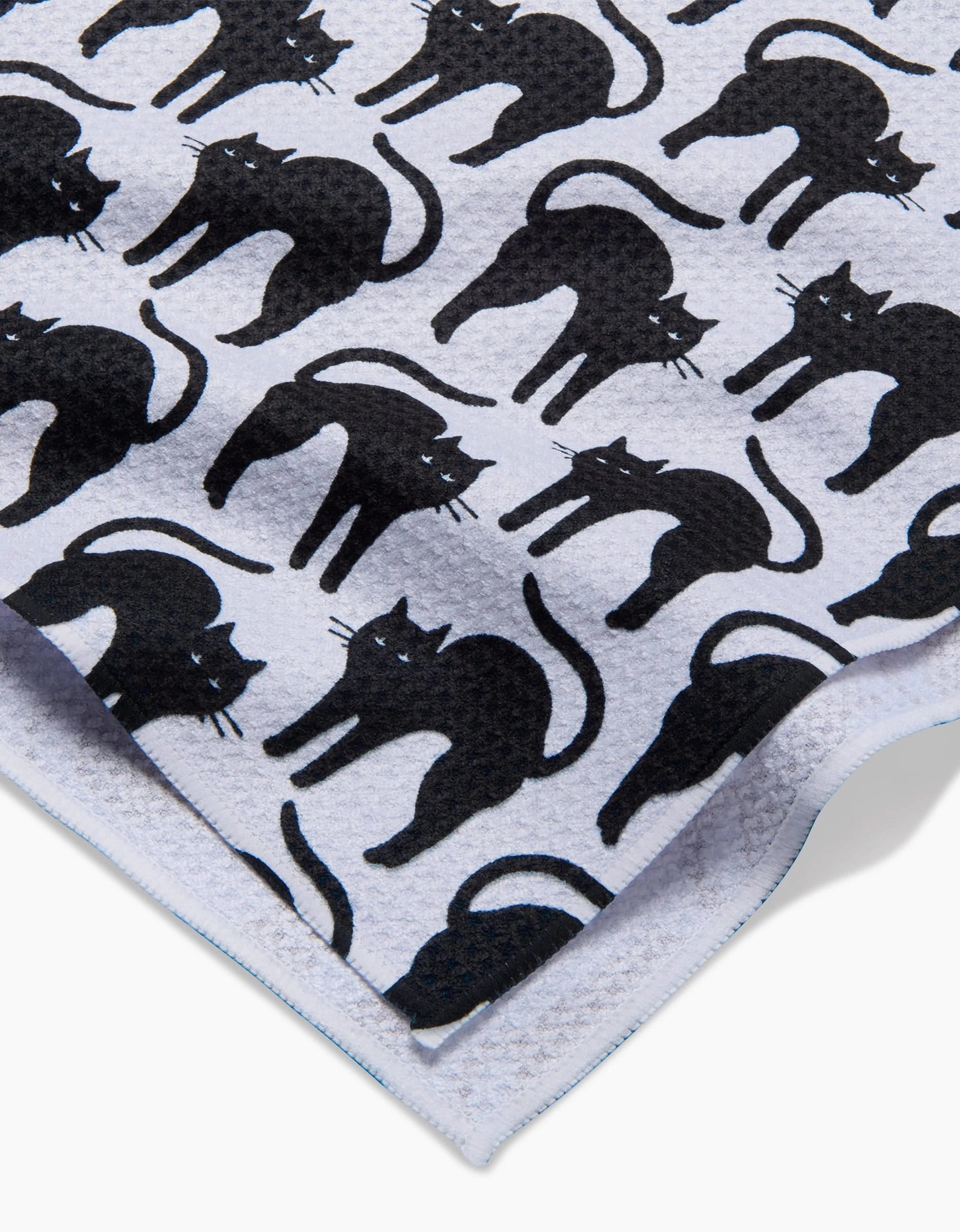 The Scaredy Cat Black tea towel by Hufton Studio features a repeating pattern of black cats on white, crafted from quick-drying recycled materials. The towel is shown slightly folded at the corner.