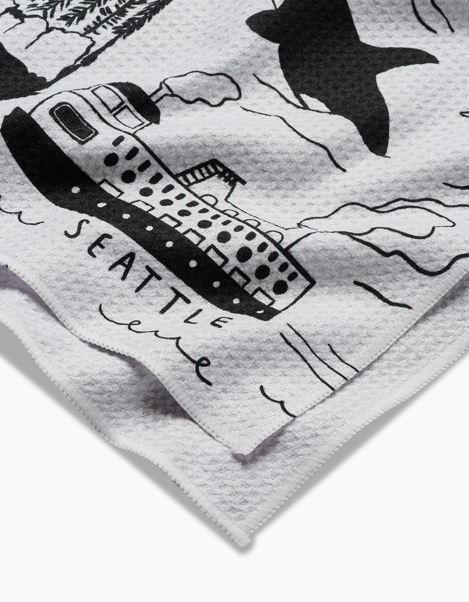A close-up of Elisabeth Wing’s Seattle tea towel, featuring white textured Geoweave fabric with black illustrations of a ferry, whale, and waves, plus “SEATTLE” written below the ferry.