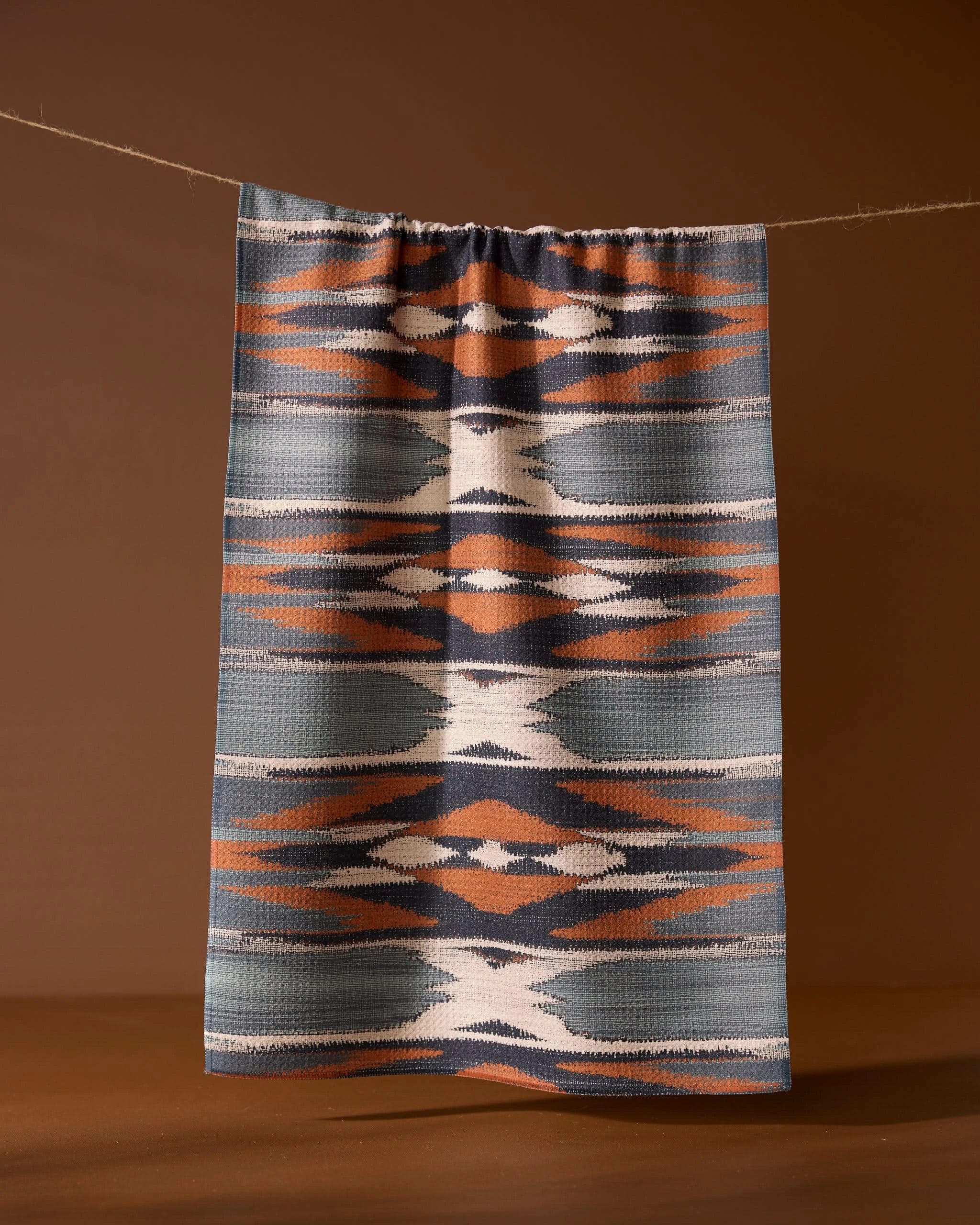 The Sedona kitchen towel by Geometry, featuring geometric blue, orange, white, and gray patterns, hangs from a string against a brown background.
