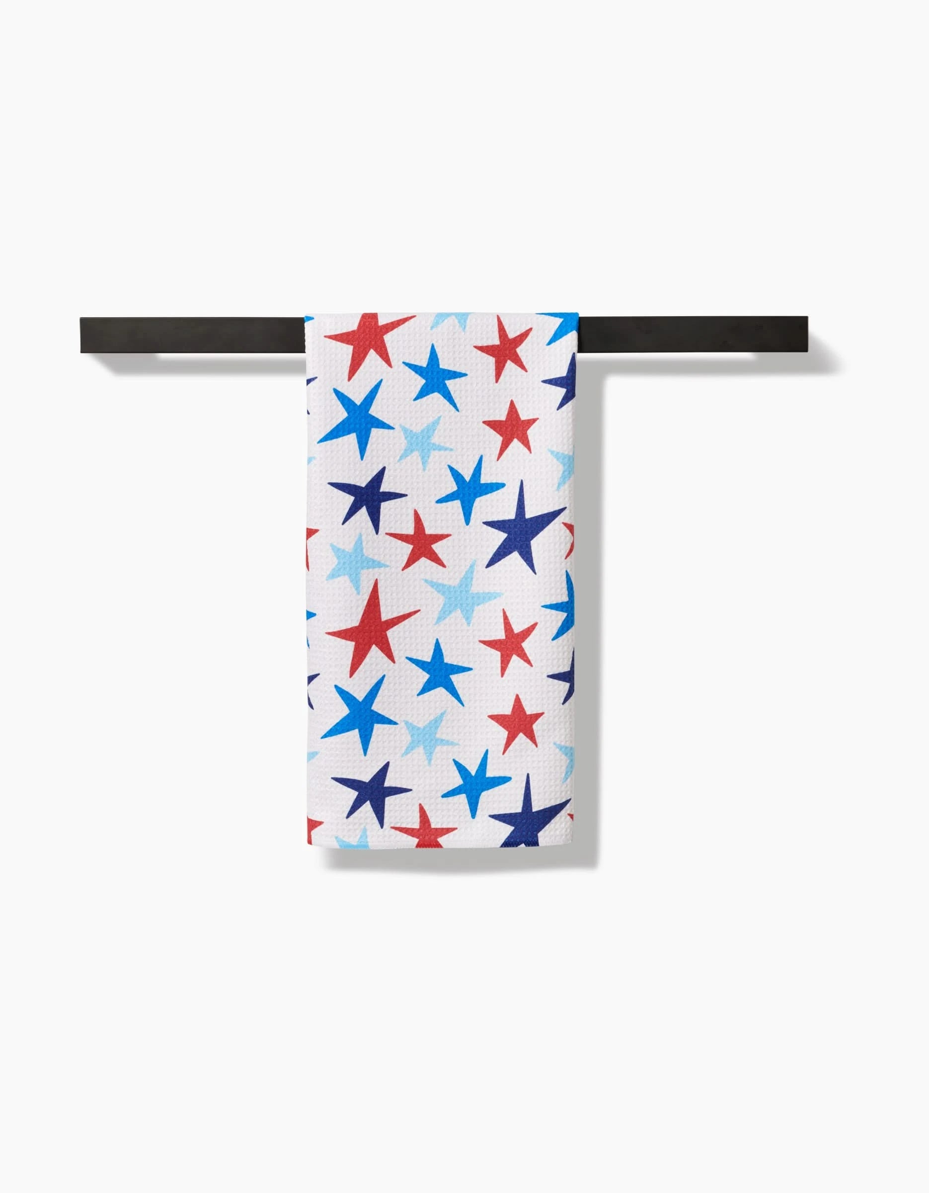 The Mindfulnice Seeing Stars tea towel, featuring red, blue, and light blue stars on white Geoweave™ fabric for premium absorbency, hangs neatly on a black rack against a white background.