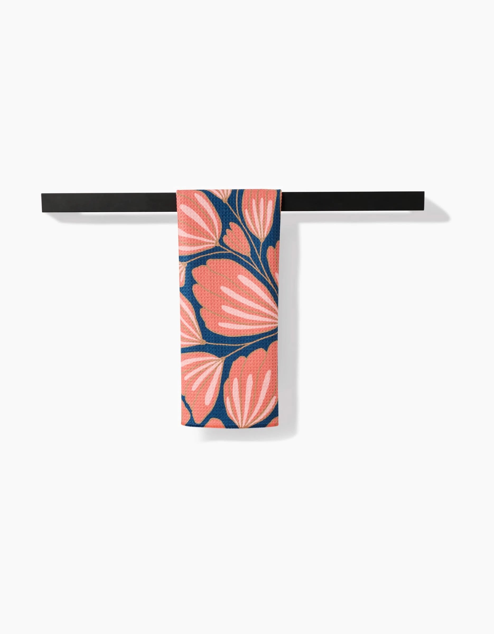 The Sheila tea towel by Amy MacCready, in coral pink with a bold navy and white floral pattern, is folded neatly over a sleek black bar against a white background.