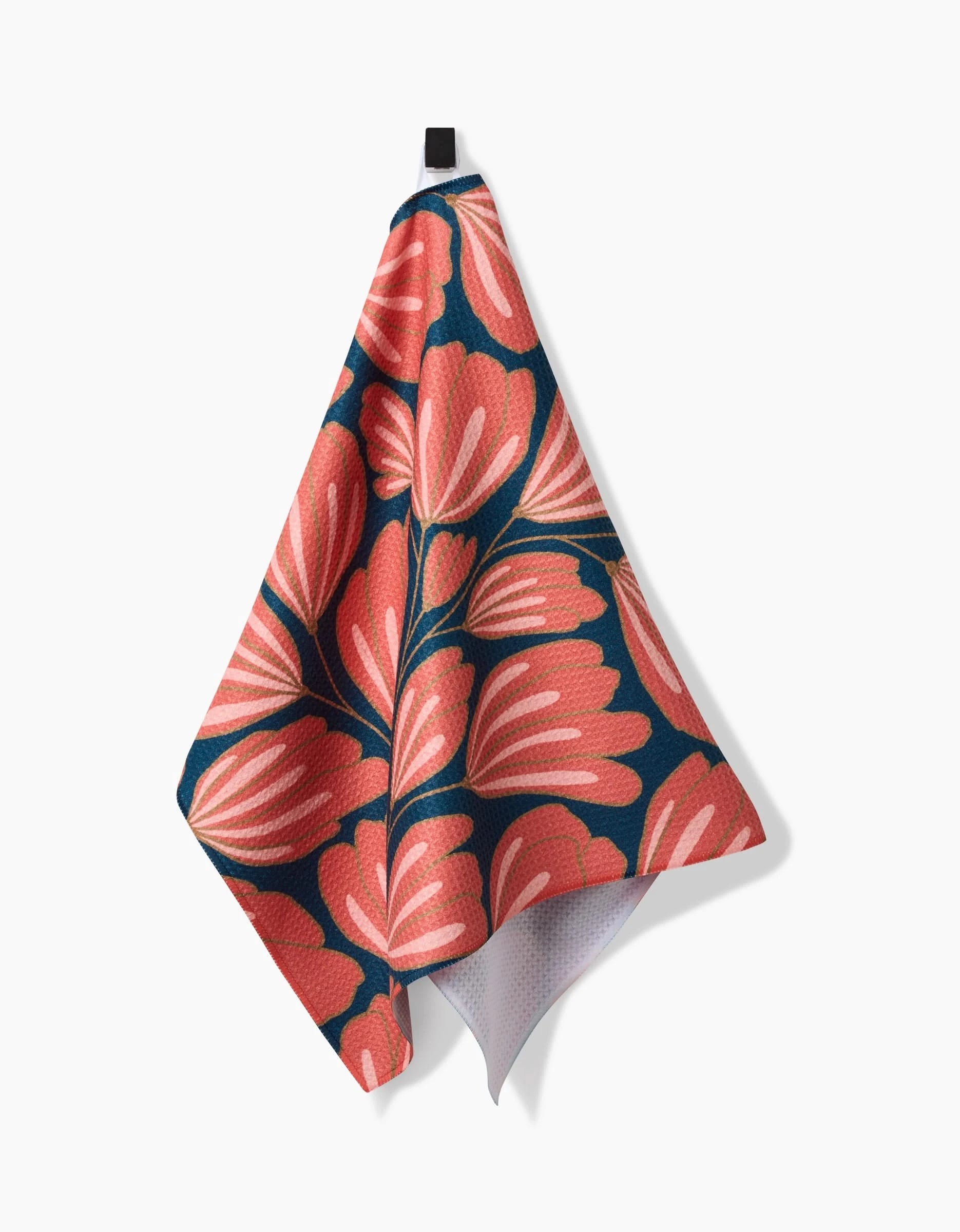 The Sheila tea towel by Amy MacCready features a bold coral pink leaf pattern on dark blue, made from ultra-absorbent recycled materials, and is shown hanging against a white surface.