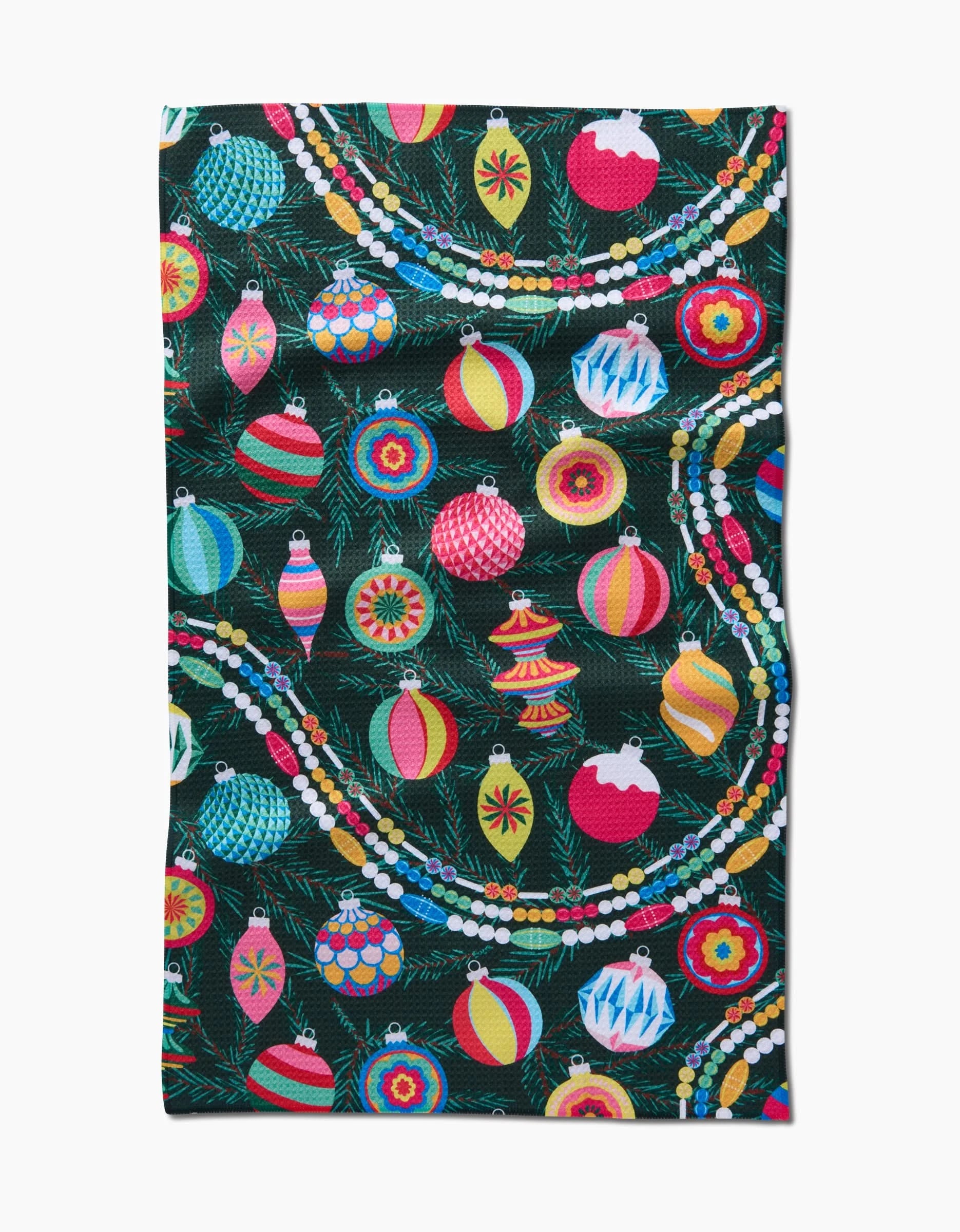 The Shiny Ornaments kitchen tea towel by Rebecca Bobko features colorful Christmas ornaments and beaded garlands on a dark green background with pine branches. Made from recycled materials, this festive towel brightens any holiday kitchen.