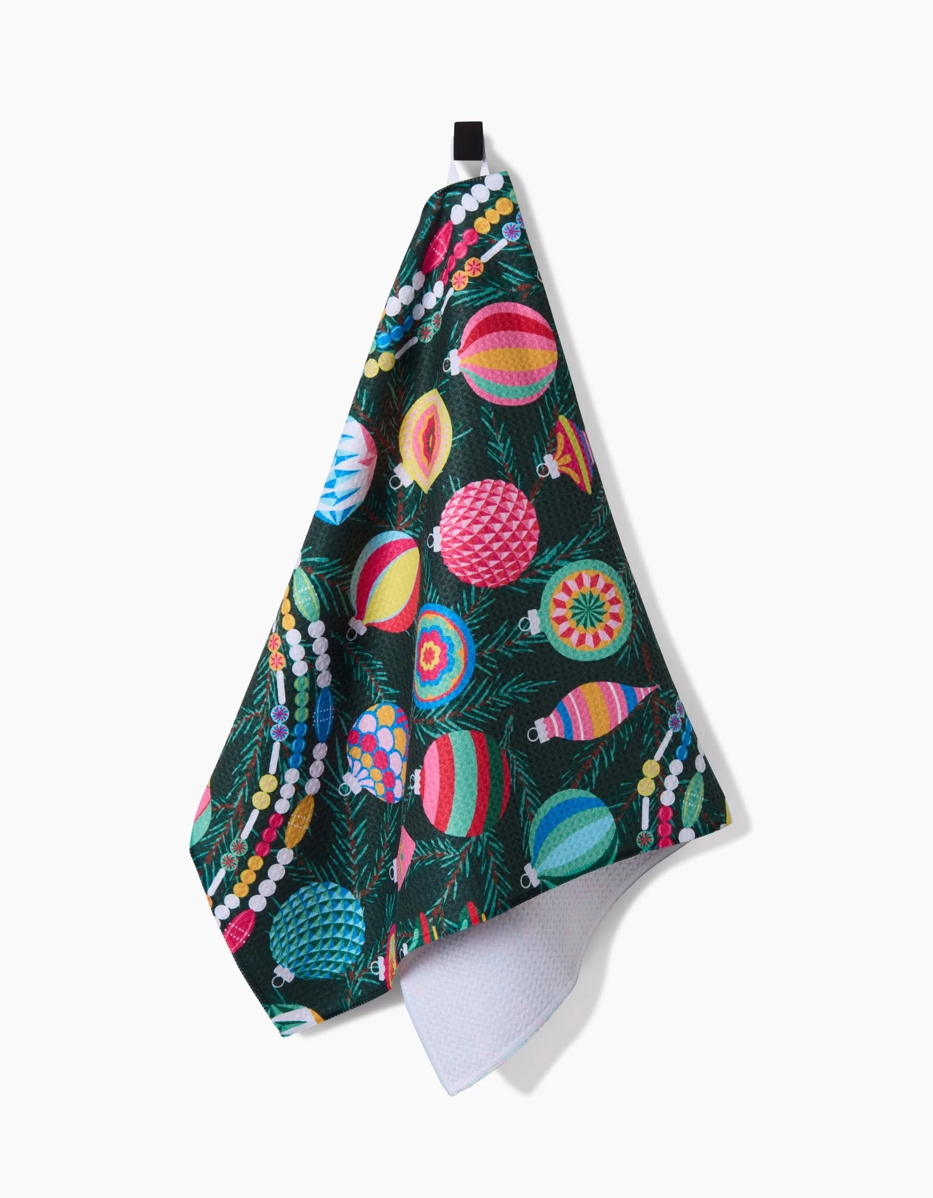 The quick-drying Shiny Ornaments towel by Rebecca Bobko hangs on a hook, displaying colorful Christmas ornaments in various shapes and stripes on a dark green, pine branch-like background.