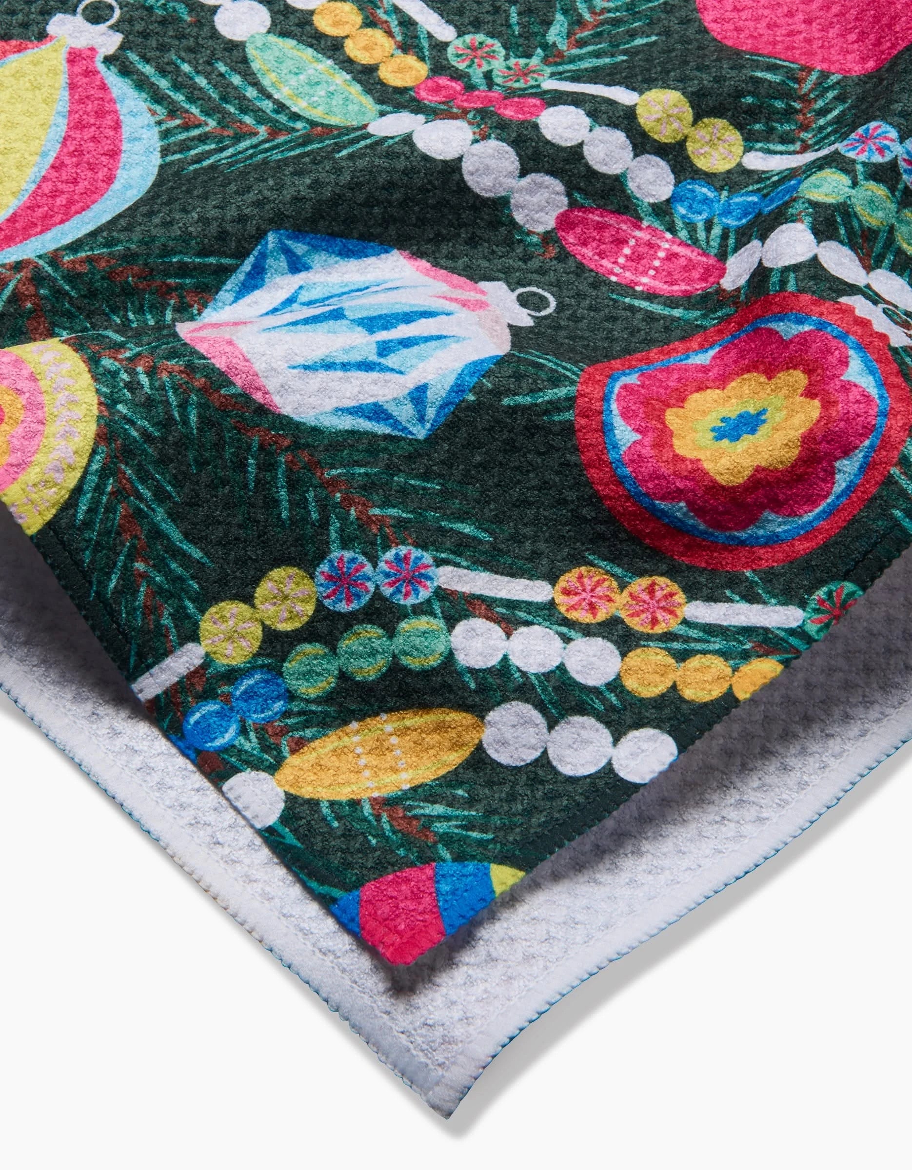 A close-up of the Shiny Ornaments tea towel by Rebecca Bobko, featuring colorful Christmas ornaments and festive patterns on a dark green, pine-needle-like background. The folded edge reveals a white underside with blue stitching.
