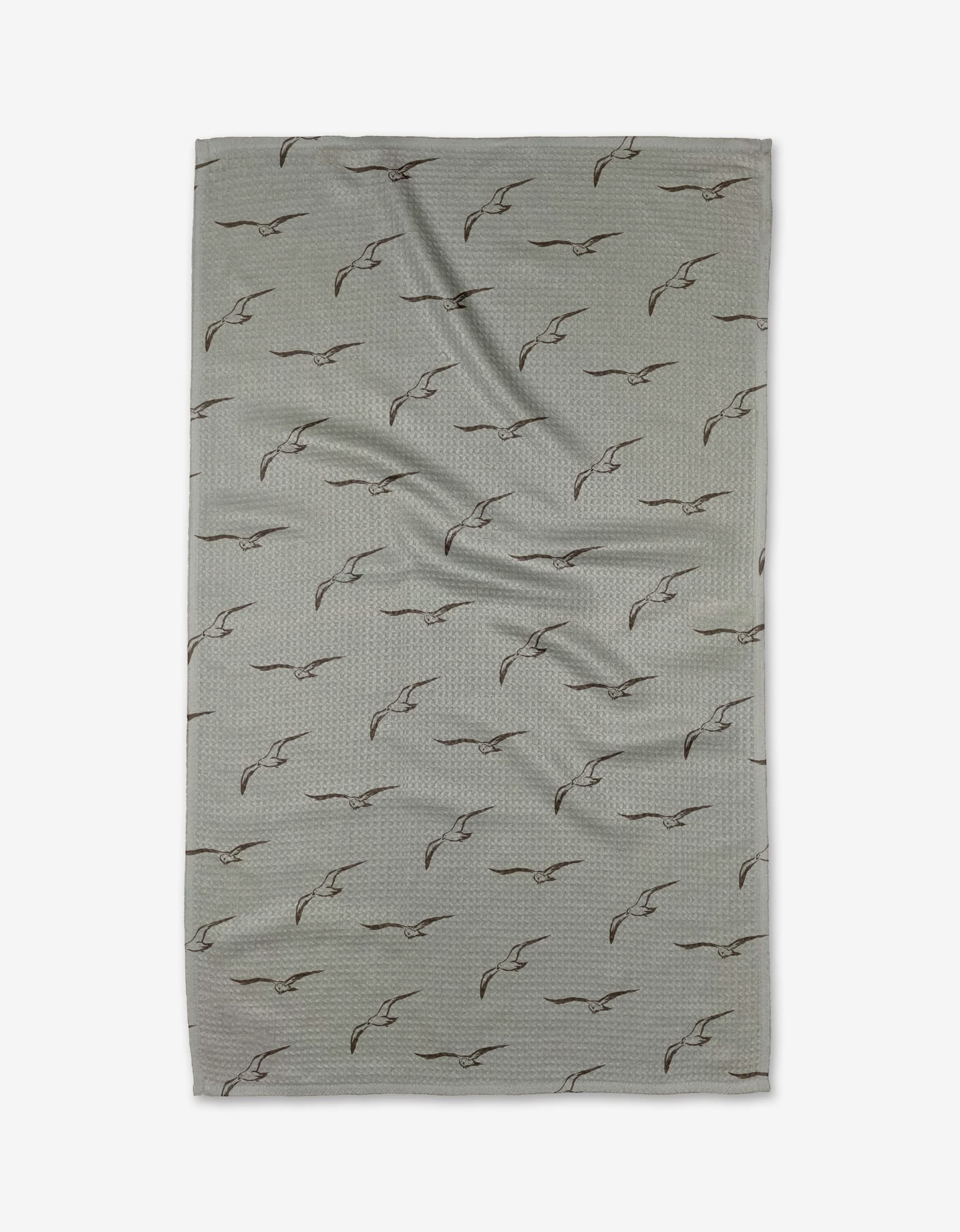 The Shoreline Scout by Jenny Keller is a light gray rectangular tea towel with black outlined flying birds in a diagonal pattern. Made from quick-drying recycled materials, it’s pictured flat with gentle creases.