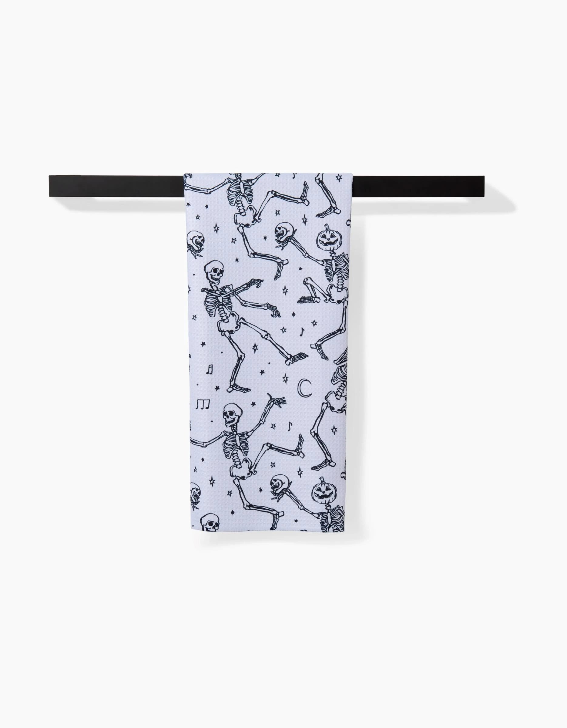 The Skeleton Halloween Dance tea towel by Aubrey Fairchild features black dancing skeletons, music notes, stars, and crescent moons on white Geoweave fabric, hanging neatly on a black towel bar against a white background.