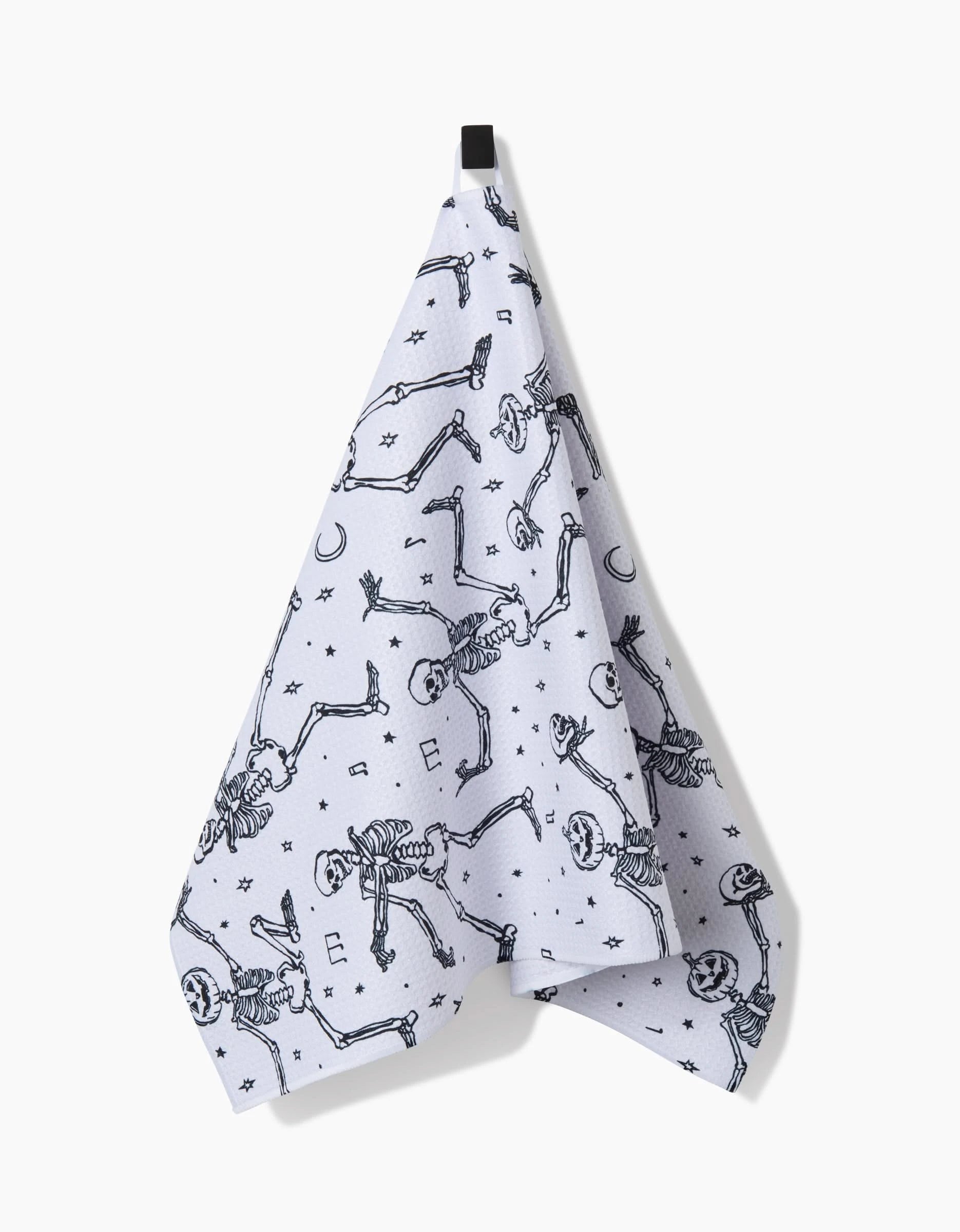 The Aubrey Fairchild Skeleton Halloween Dance white tea towel, made from recycled materials, hangs from a black hook and features playful skeletons dancing among stars and crescent moons.