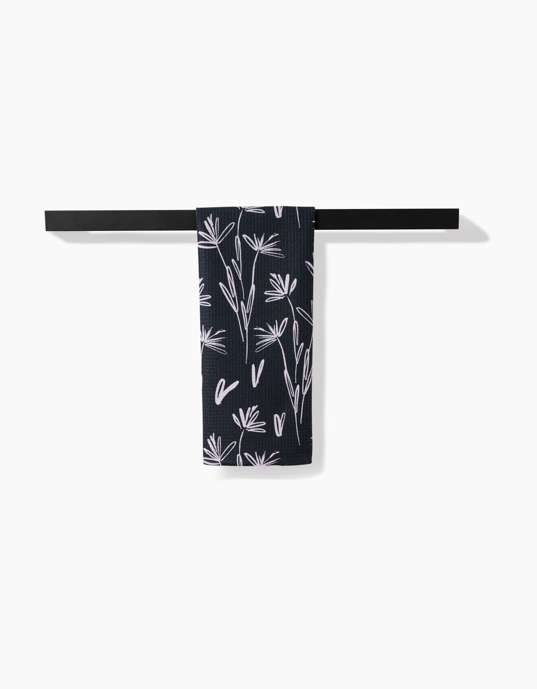 A black towel rack holds a Toboggan Avenue Sketchbook Dust tea towel, featuring dark Geoweave™ fabric with a white floral pattern, hanging neatly against a plain white background.