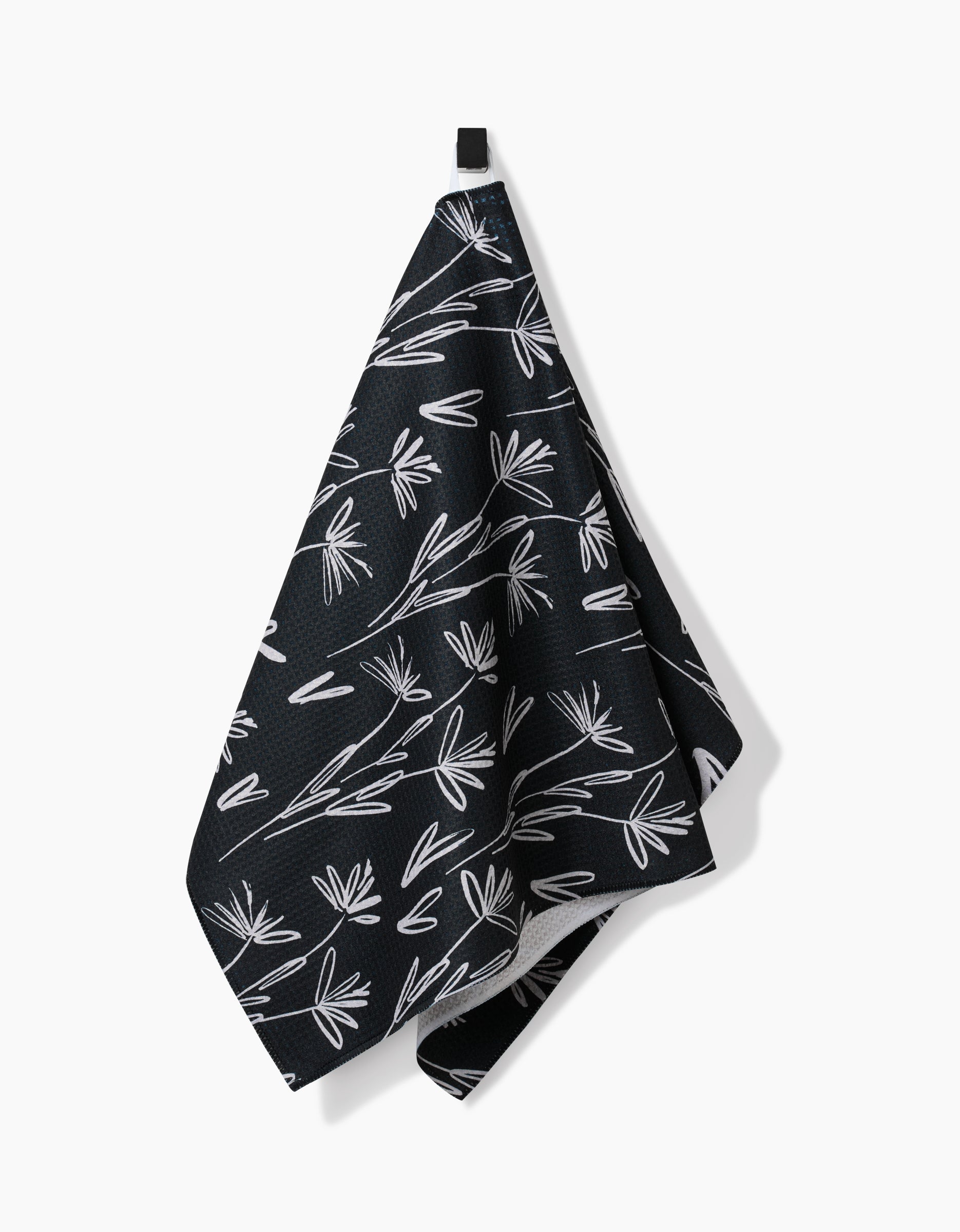The Toboggan Avenue Sketchbook Dust Tea Towel, featuring a white leaf pattern on black Geoweave™ fabric, hangs from a black hook against a plain white background.