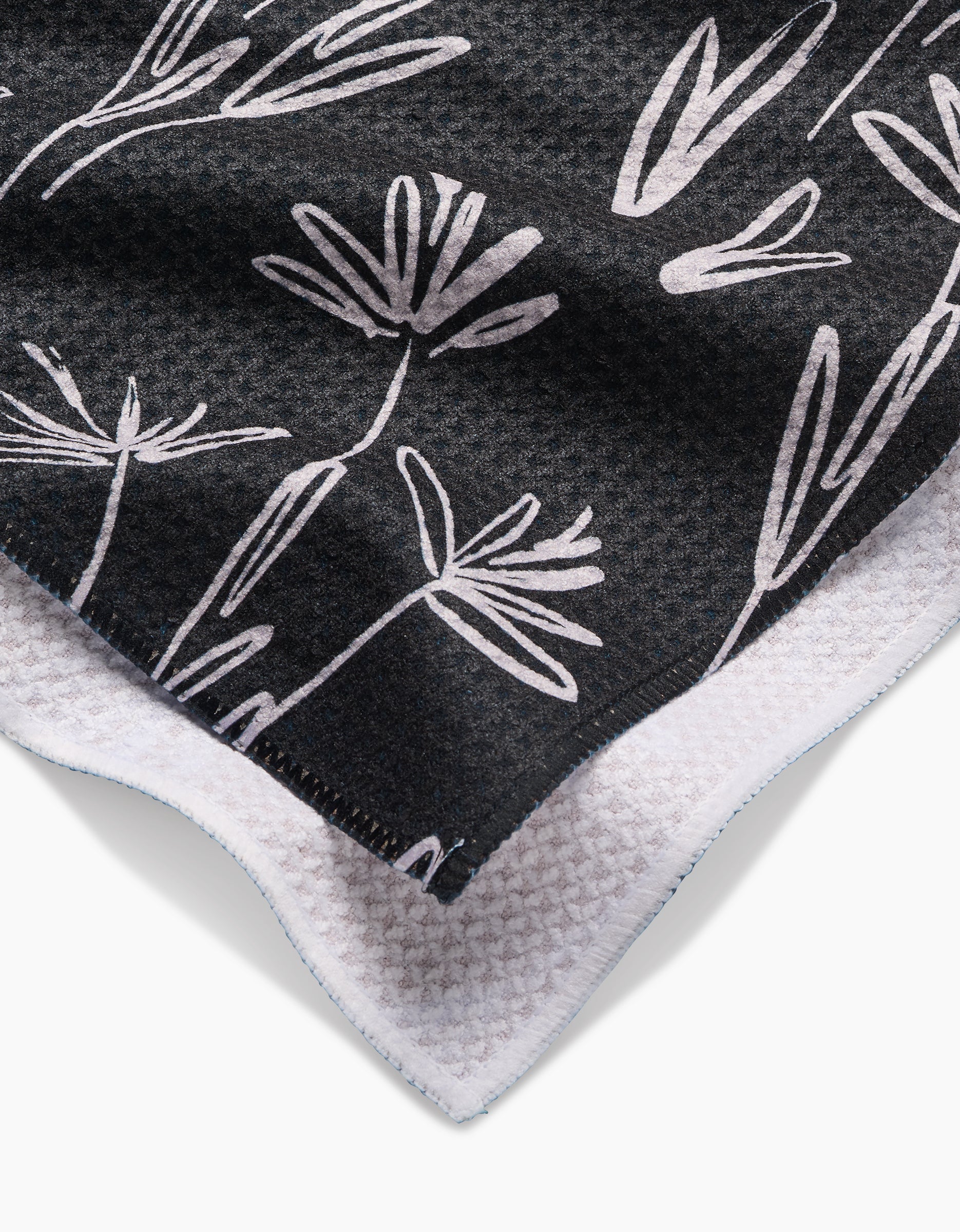 A close-up of the Toboggan Avenue Sketchbook Dust Tea Towel in black Geoweave™ fabric, displaying a white floral pattern and textured white border, with one corner folded over.