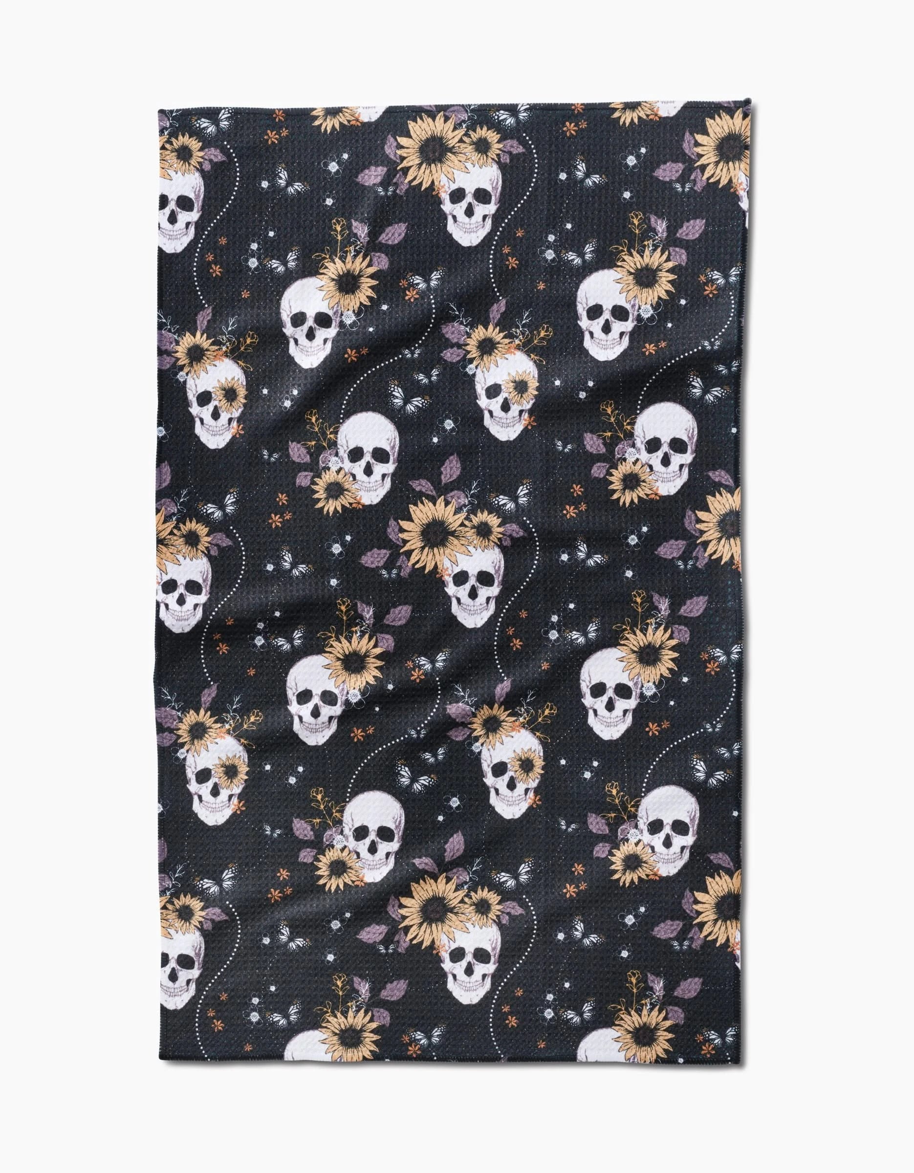 The Skulls In Bloom tea towel by Ashley Celena features a dark background with white skulls, yellow sunflowers, orange stars, and purple leaves in a repeating pattern across its textured surface.