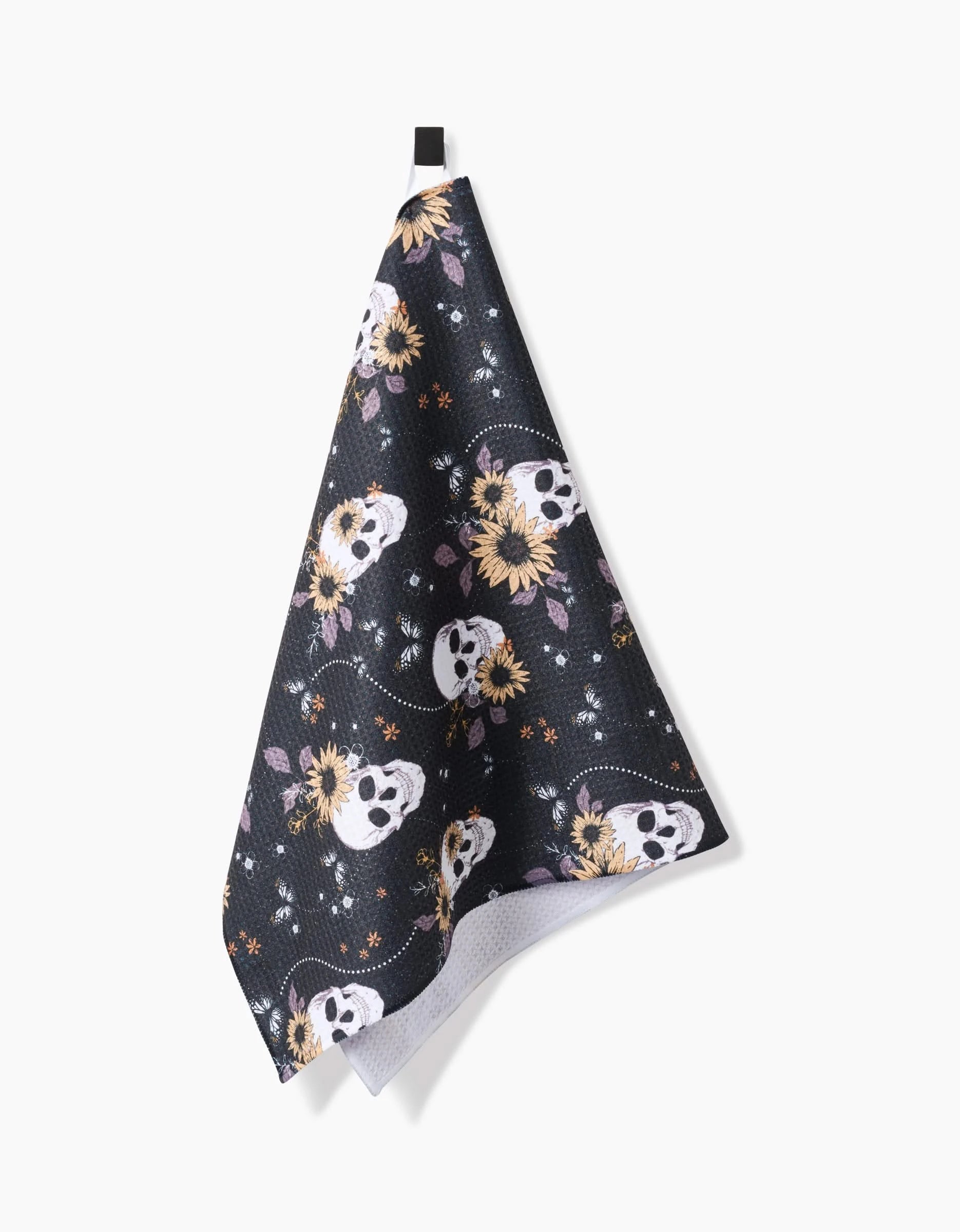 The Ashley Celena Skulls In Bloom tea towel features a quick-drying black fabric with white sugar skulls, sunflowers, purple leaves, and decorative dots, and hangs from a black loop.