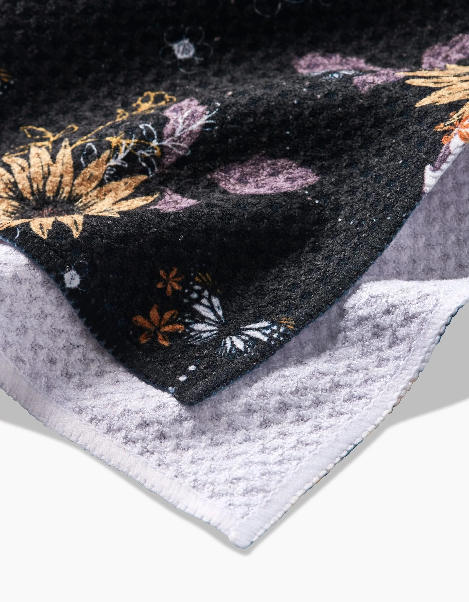 A close-up of Skulls In Bloom by Ashley Celena—a textured black fabric with colorful floral and butterfly patterns—layered over a light gray, quick-drying waffle-knit tea towel.