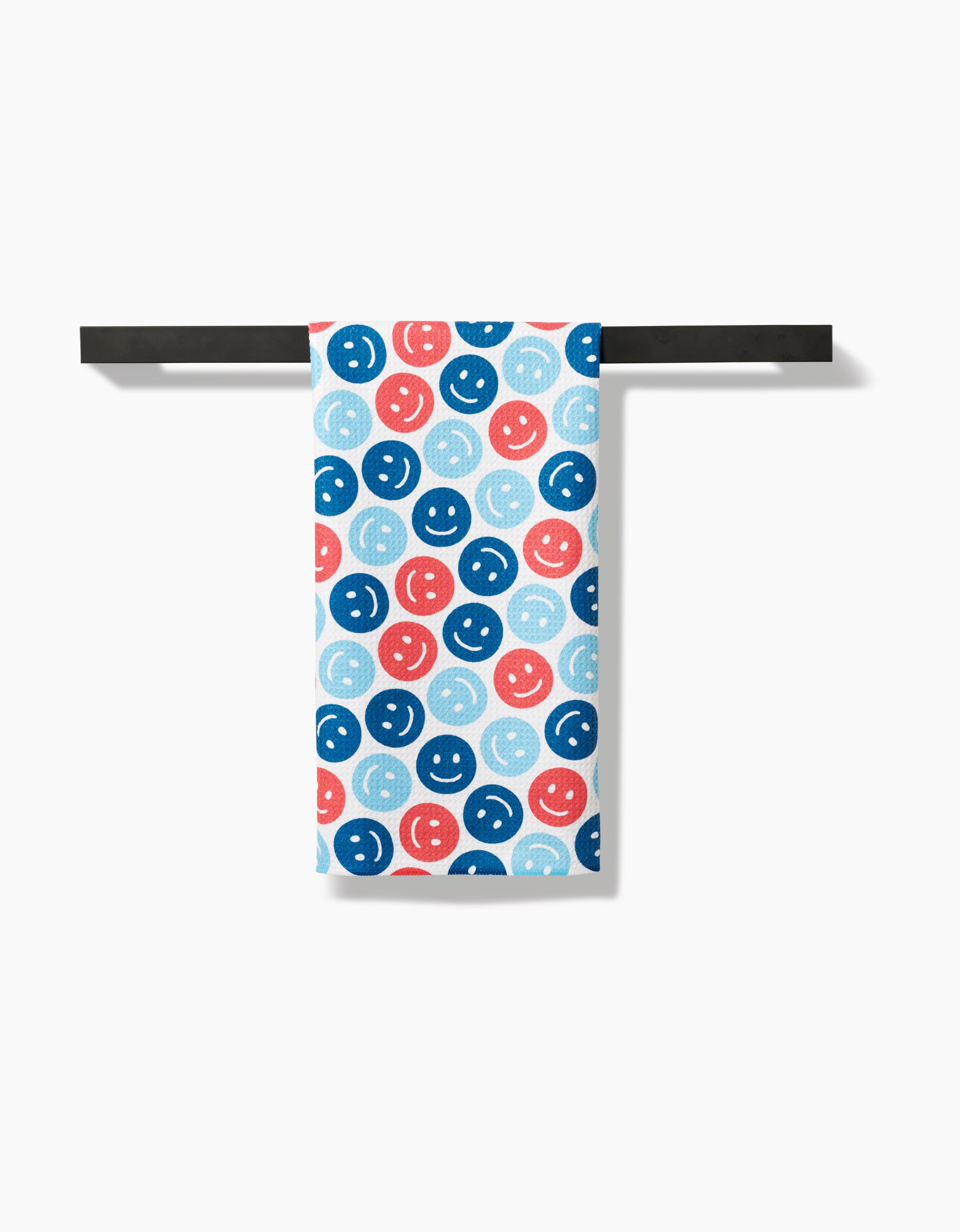 The Hufton Studio Smiley Face July tea towel, featuring blue, red, and light blue smiley faces on white recycled material, hangs on a black bar against a plain white background.