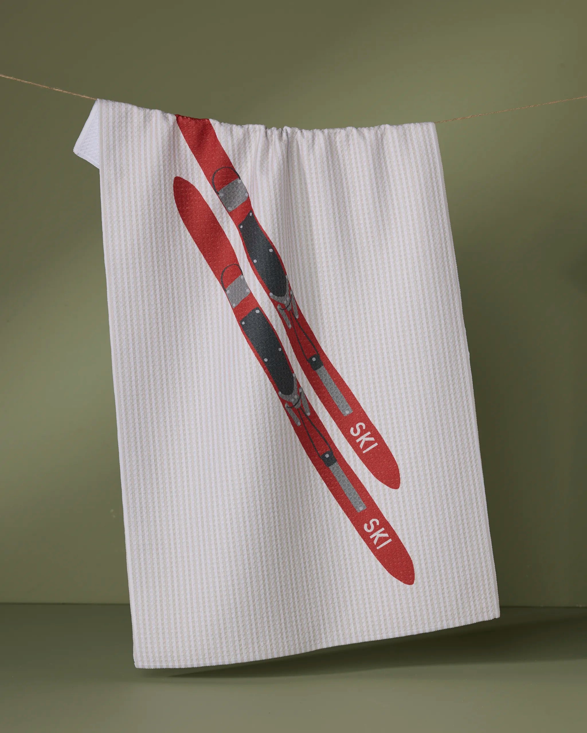 The "Snowcap" tea towel by Geometry hangs on a line, featuring subtle vertical stripes and an illustration of two red skis with black bindings and "SKI" near the tips. Made from ultra-absorbent recycled materials.