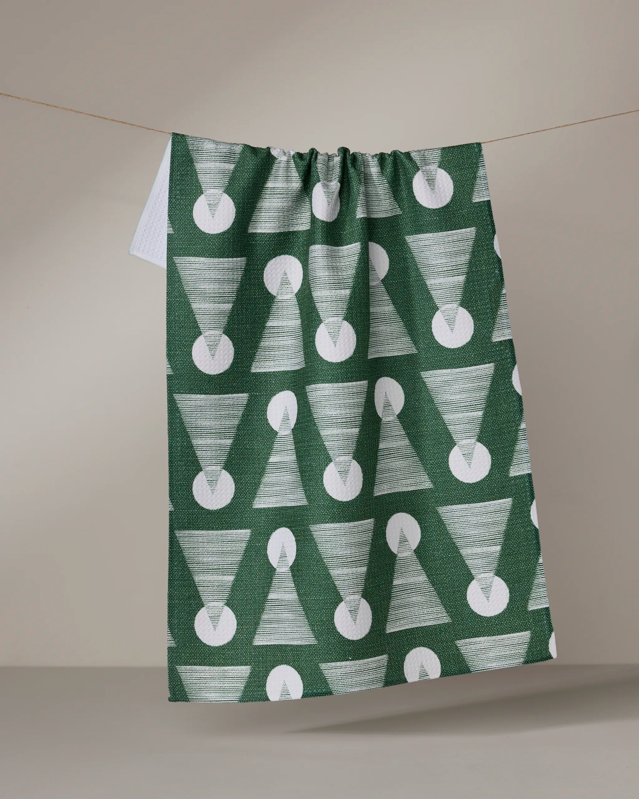 The Geometry Soltice Glow kitchen towel, made from recycled materials and featuring a green design with repeating white candle shapes, hangs from a clothesline against a plain, neutral background.