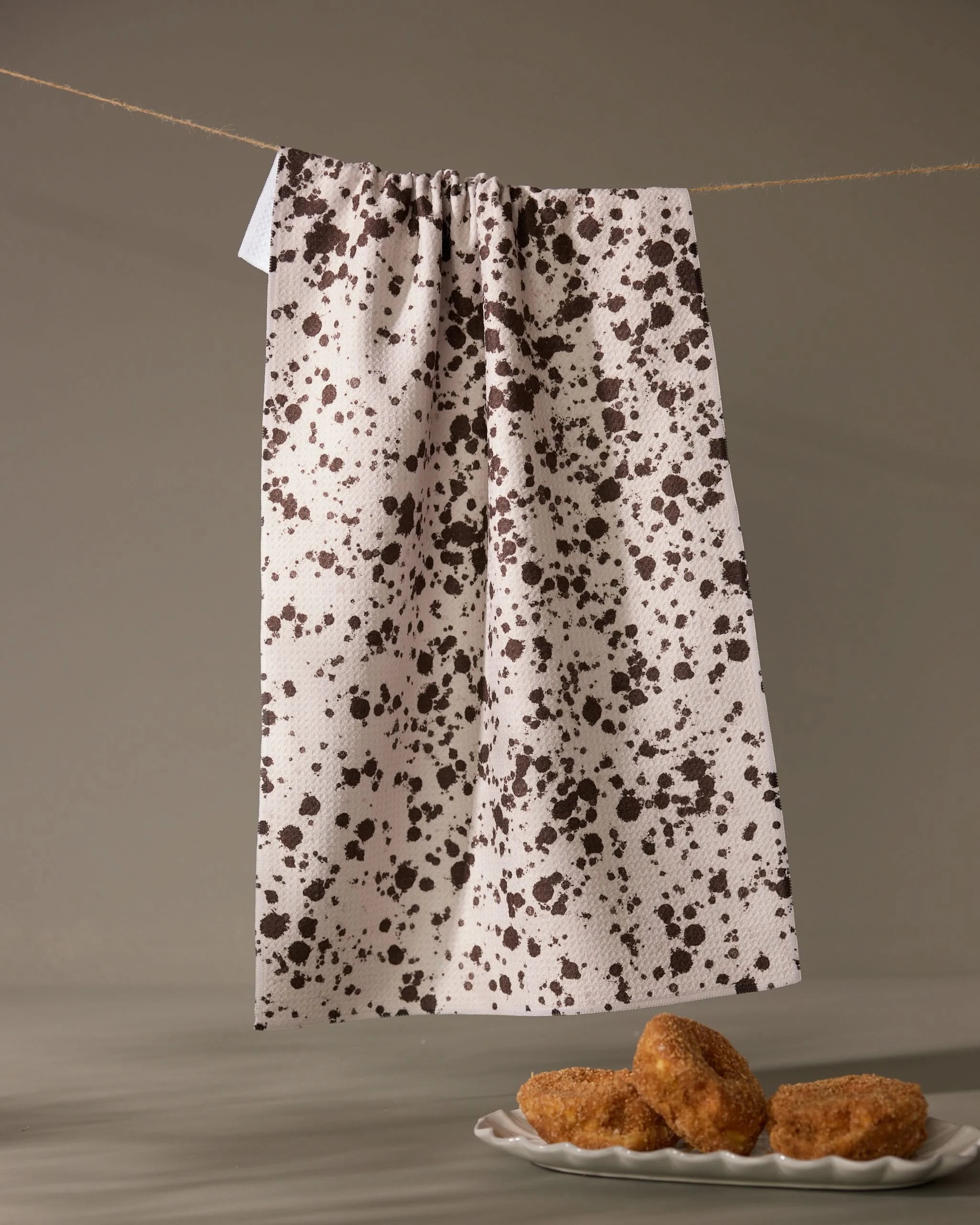 The Geometry Spill white absorbent dish towel with black speckles hangs on a string, as a plate with four sugar-coated pastries rests on a light gray surface below.