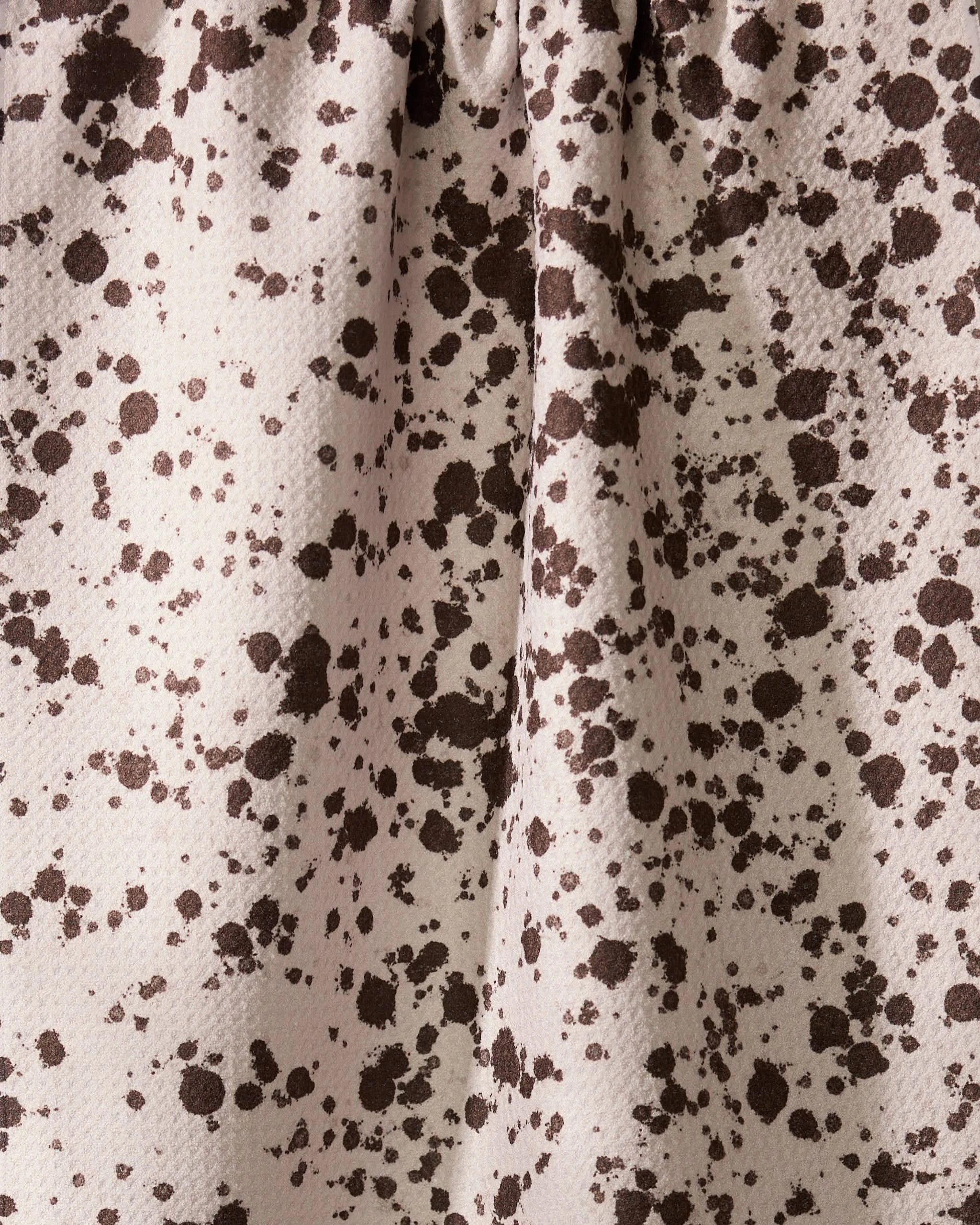 The Geometry Spill dish towel features beige fabric with dark brown spots and splatters, resembling paint stains. Its absorbent surface has a visible texture and gentle folds.