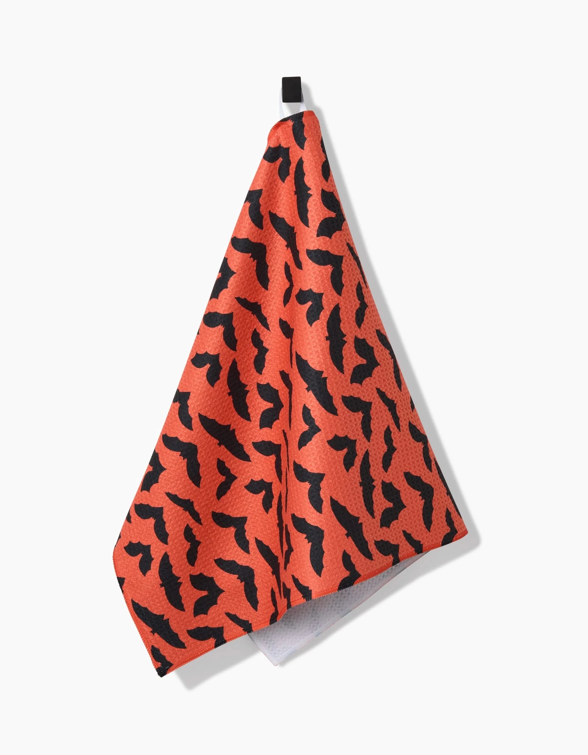 The Spooky Bats tea towel by Rebecca Bobko, featuring a black abstract bat pattern, hangs from a small black loop against a white background.