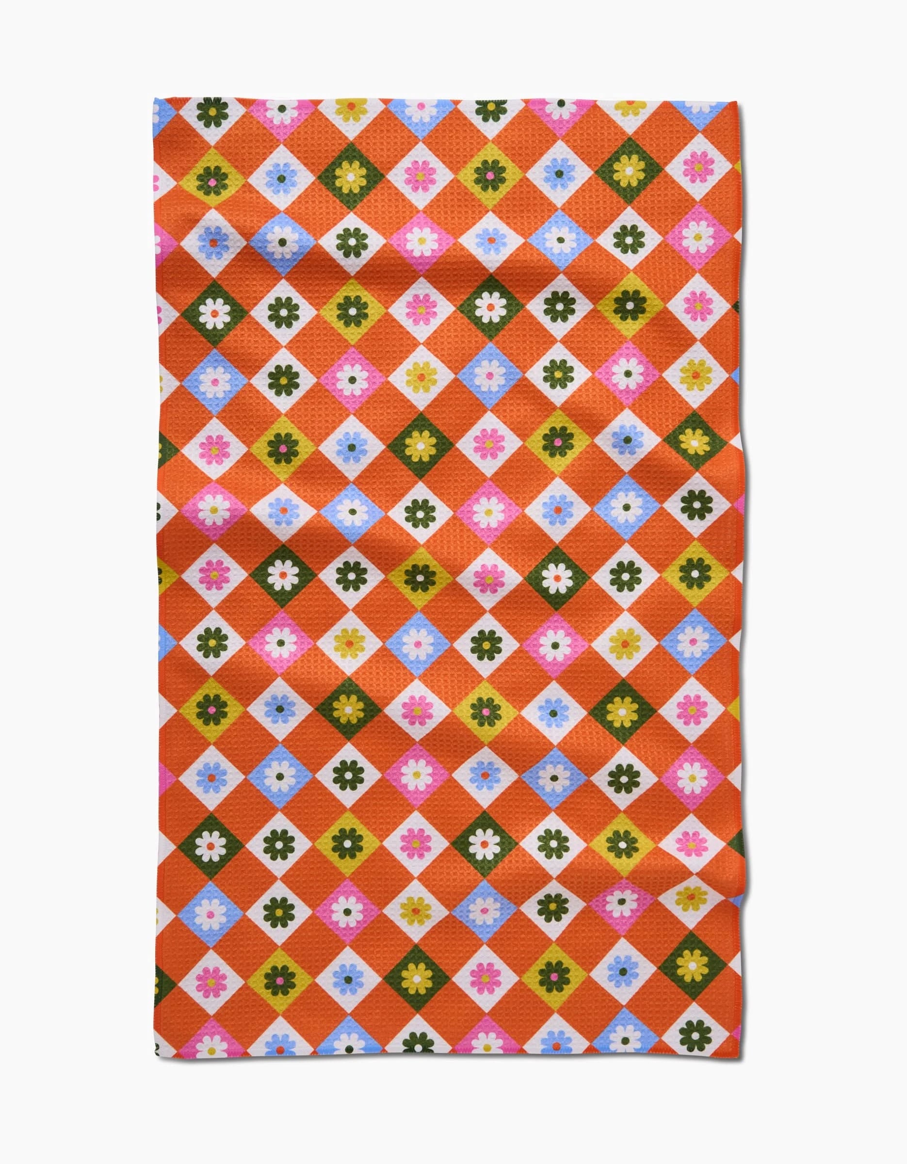 The Spring Check tea towel by Megan Roy features a rectangular Geoweave fabric with colorful diamond patterns in orange, green, blue, pink, and white, each adorned with a small central flower motif. The fabric appears slightly wrinkled.