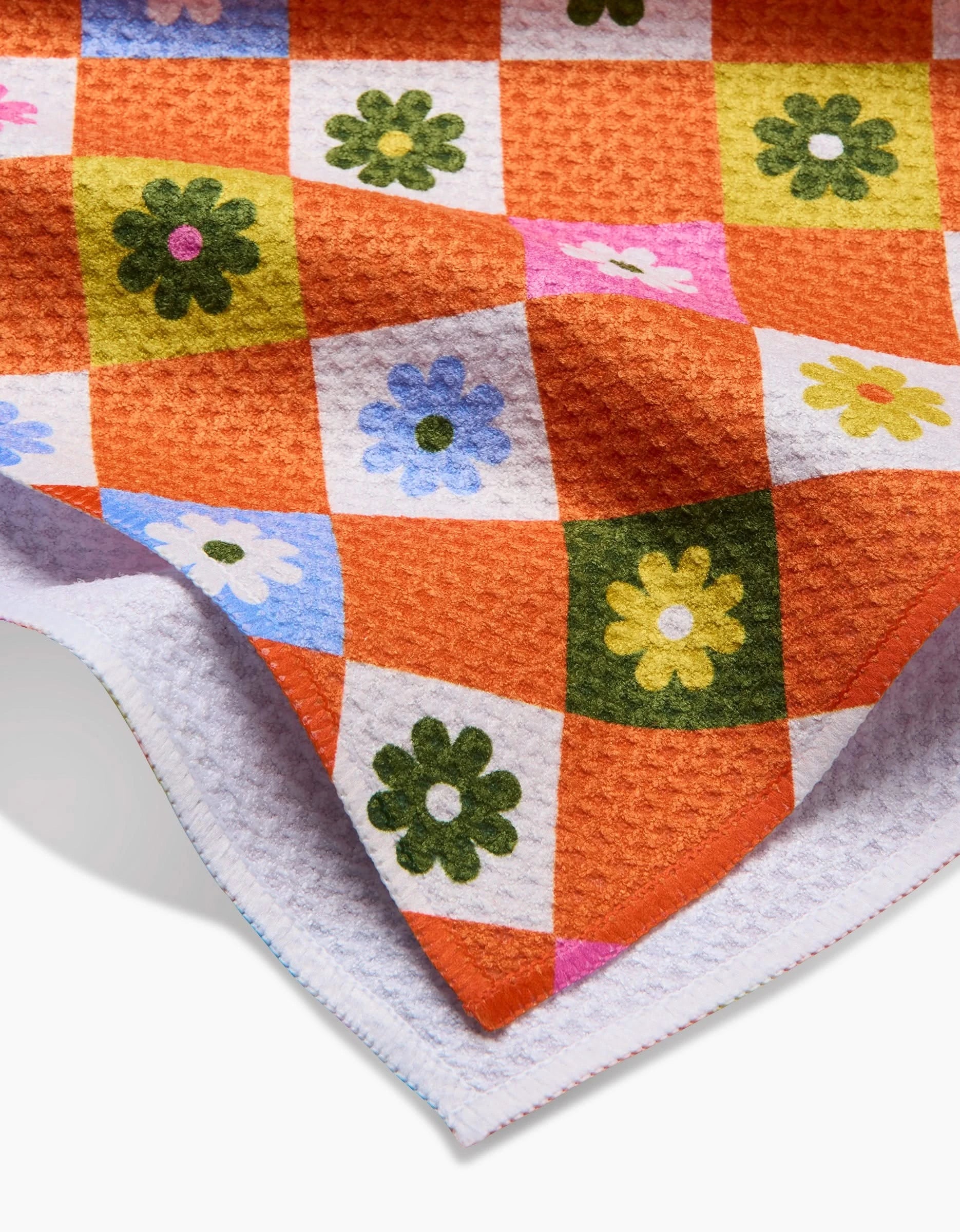 A close-up of the Megan Roy Spring Check tea towel shows its textured Geoweave fabric with orange and white checkered pattern and colorful flower designs, softly folded at the corner.