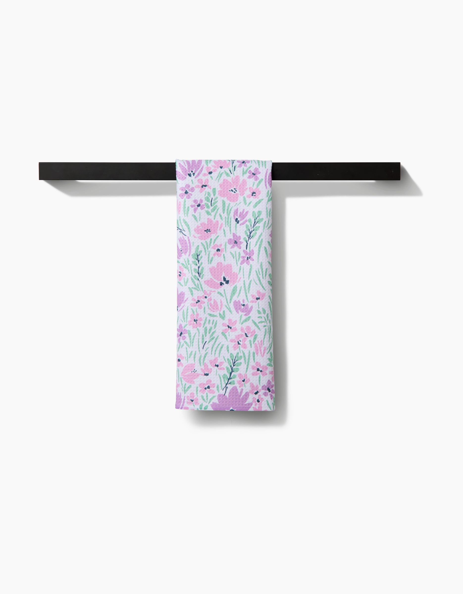 The Spring Fleur Blooms tea towel by Hufton Studio, featuring a pink and green floral design on recycled fabric, hangs neatly over a black horizontal towel rack against a white background.