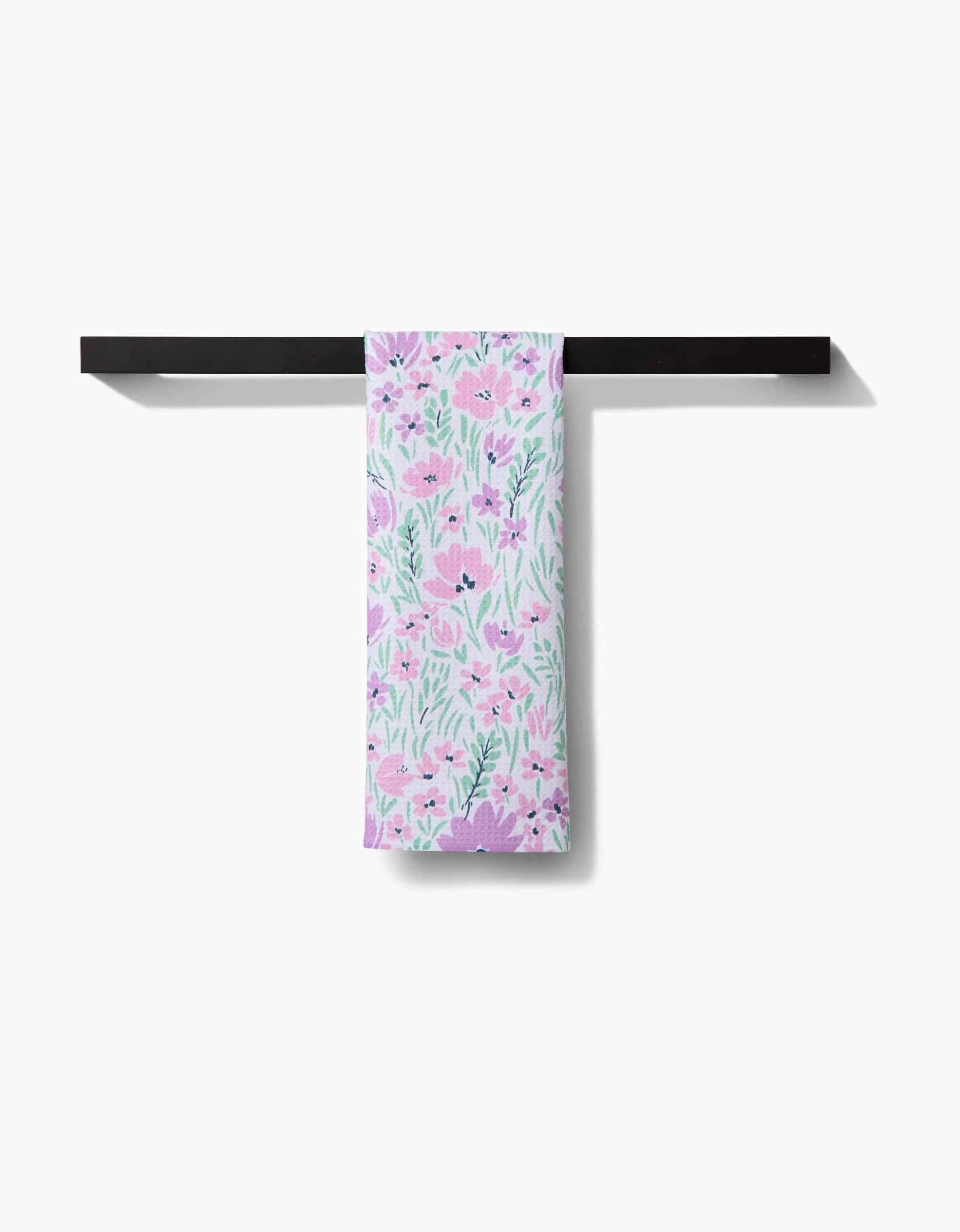 The Spring Fleur Blooms tea towel by Hufton Studio, featuring a pink and green floral design on recycled fabric, hangs neatly over a black horizontal towel rack against a white background.