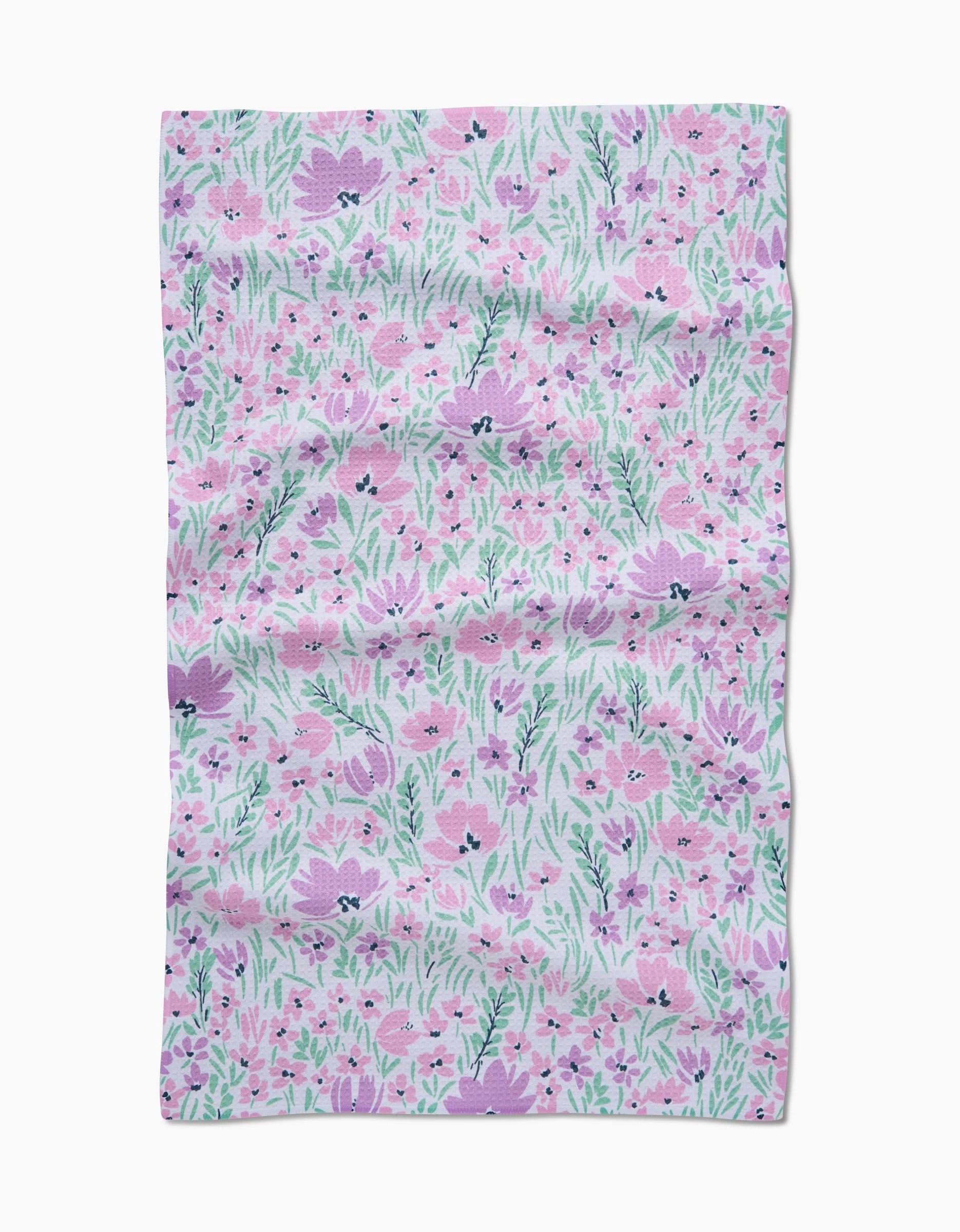 The Hufton Studio Spring Fleur Blooms Geoweave™ tea towel features a floral design with pink and purple flowers and green leaves on a light background, displayed with visible creases and wrinkles.
