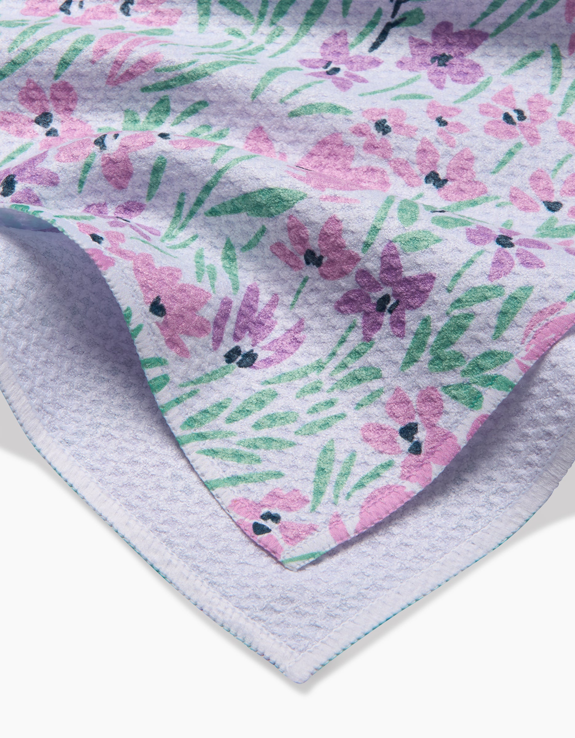 A close-up of the Hufton Studio Spring Fleur Blooms tea towel, showing pink and purple floral patterns on a light background with a textured underside made from recycled materials.