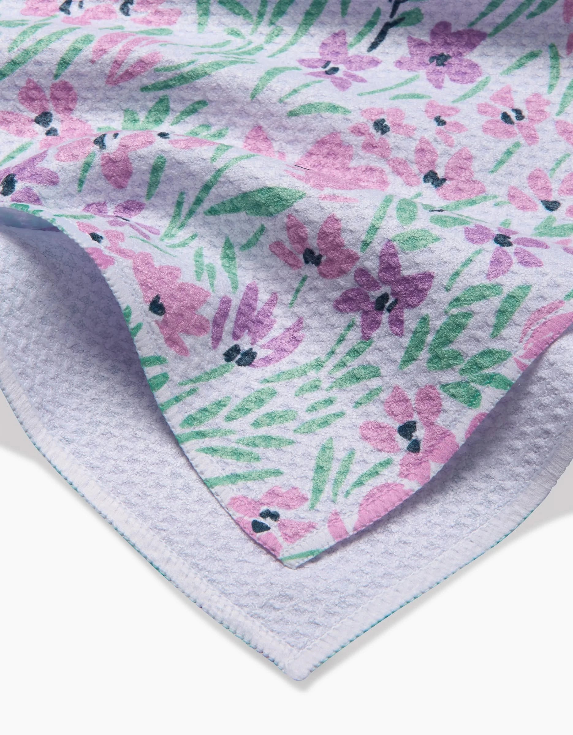 A close-up of the Hufton Studio Spring Fleur Blooms tea towel, showing pink and purple floral patterns on a light background with a textured underside made from recycled materials.